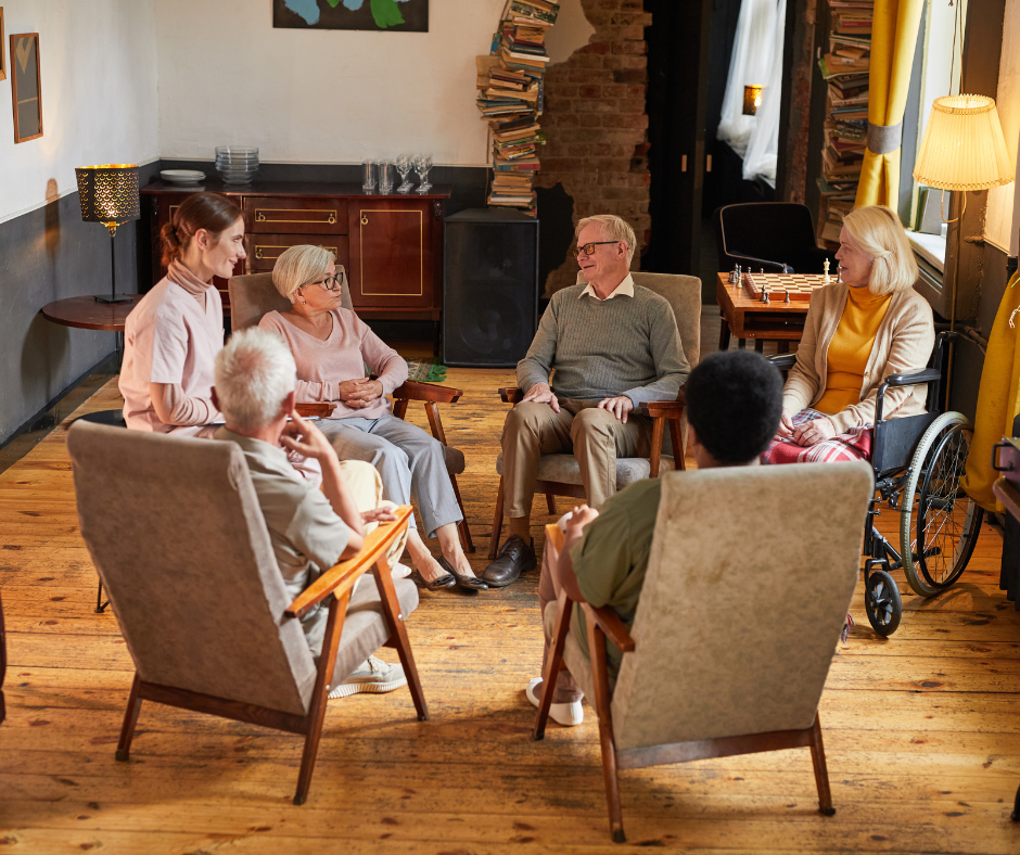 Group of older adults and a caregiver in chairs, engaged in a conversation in a room.
