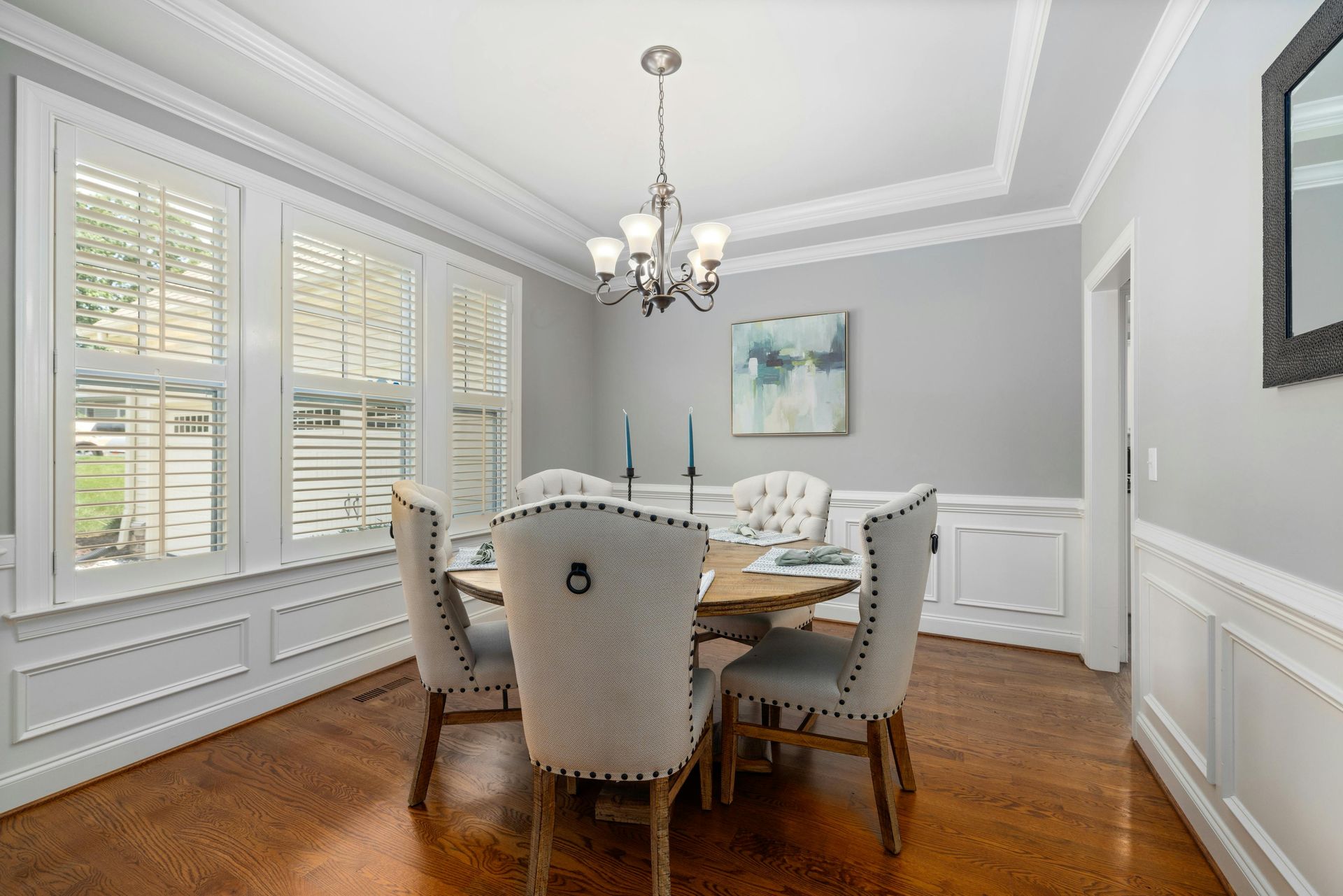 Dining room with round table, six chairs, chandelier, and artwork. Hardwood floors and white trim.
