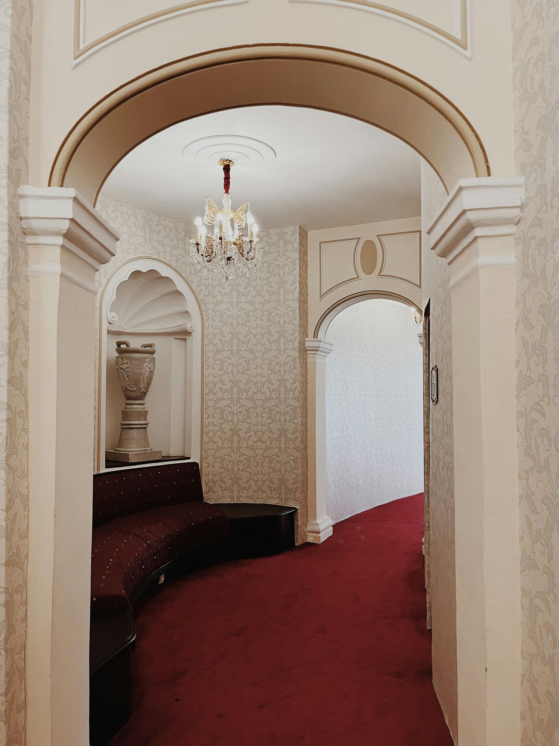 Arched doorway leading to a hallway with a red carpet and a decorative alcove with a sculpted vase and bench.