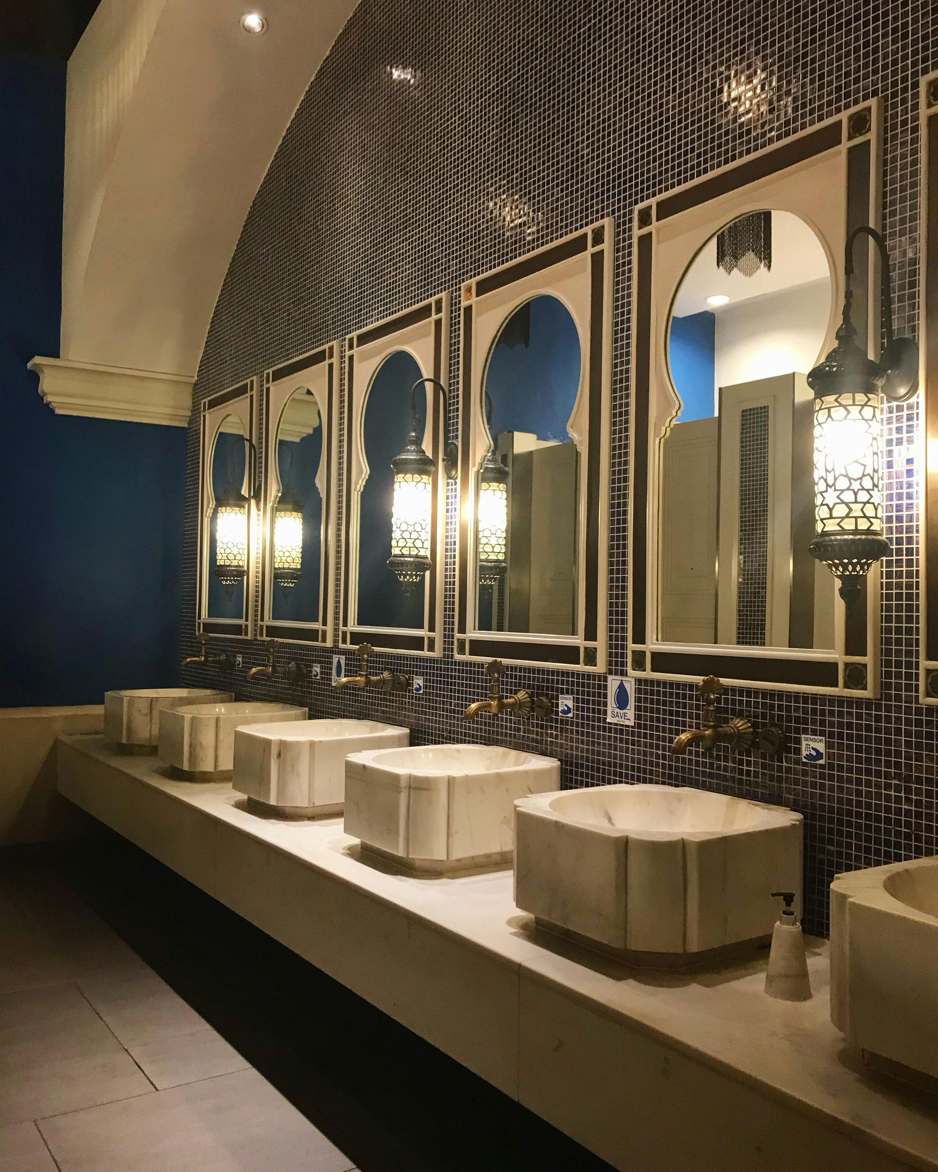 Public restroom with ornate mirrors and sinks. Dark blue and white tile. Arched mirrors with lanterns.