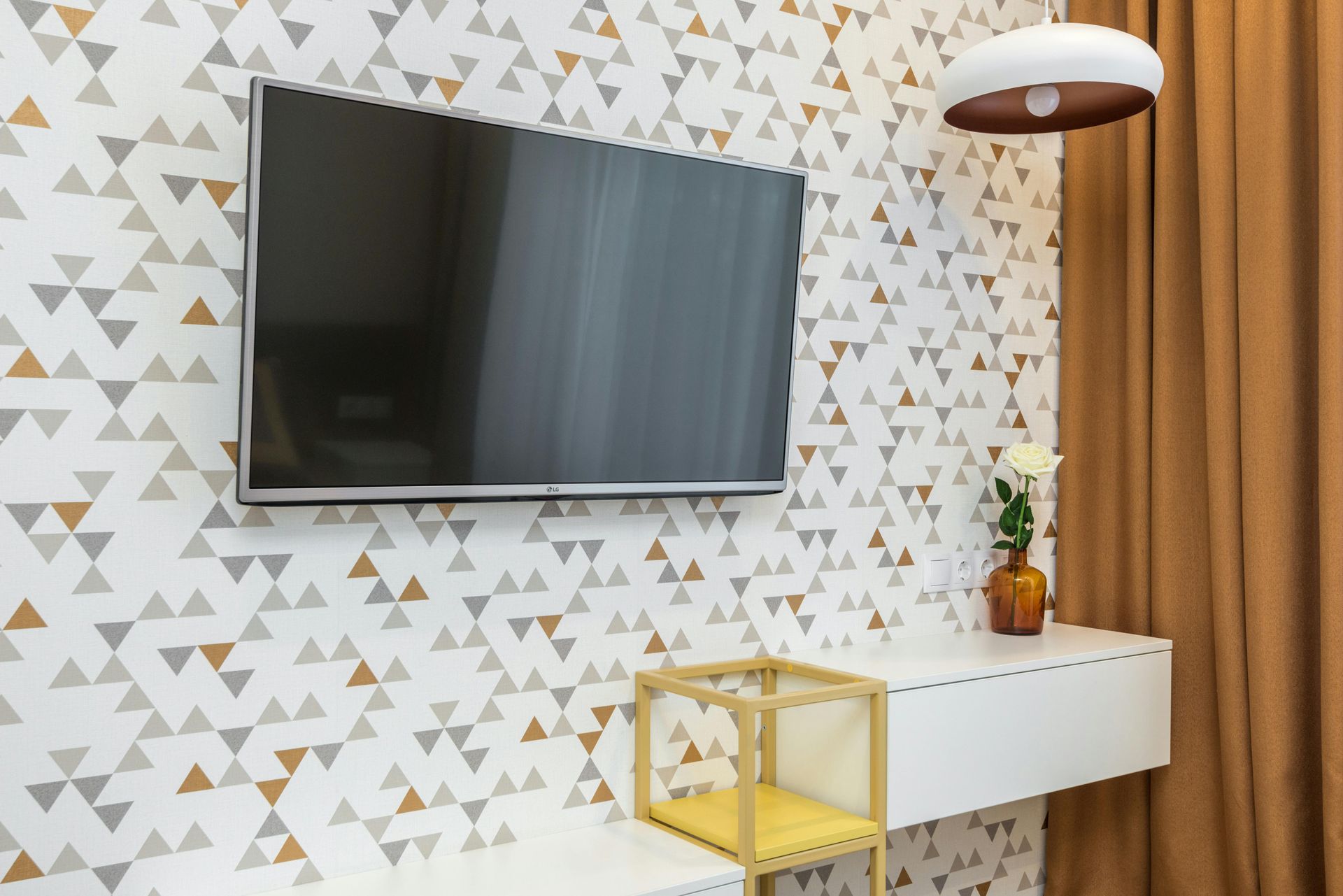 TV mounted on patterned wallpaper above a white floating shelf, with gold accents and brown curtain.