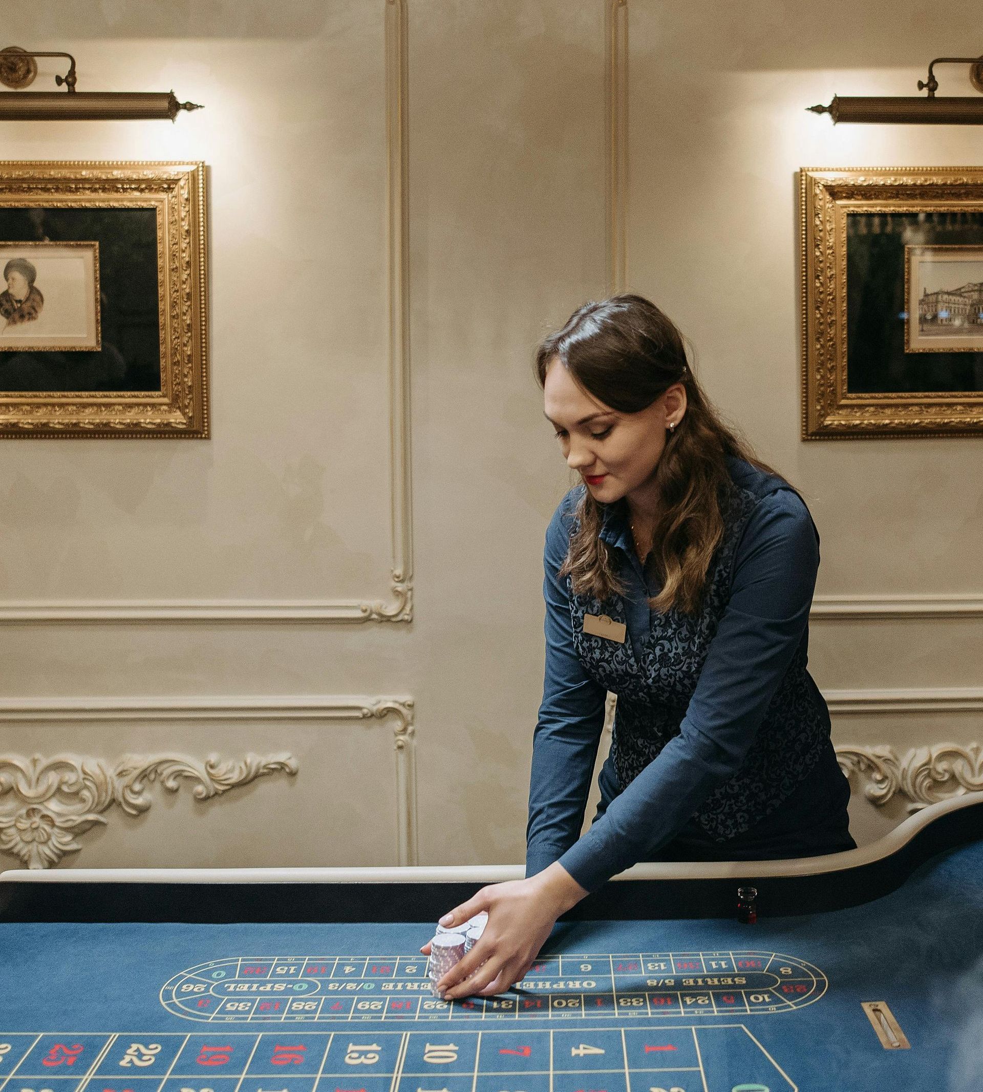 Woman in a casino uniform handling cards at a roulette table. Ornate wall decor and lighting.