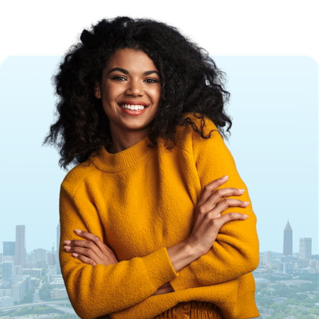 Woman with arms crossed, smiling broadly, wearing a yellow sweater; city background.
