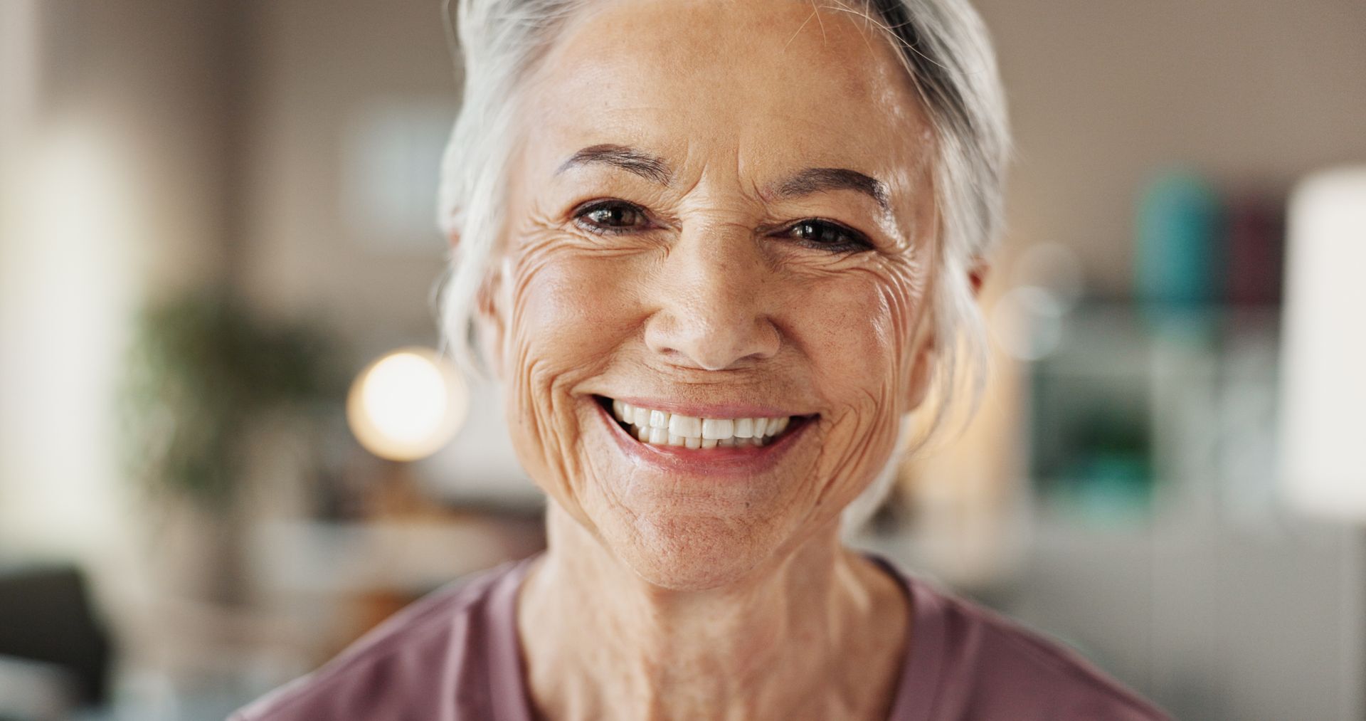 Smiling person in a softly lit home interior, wearing a mauve top