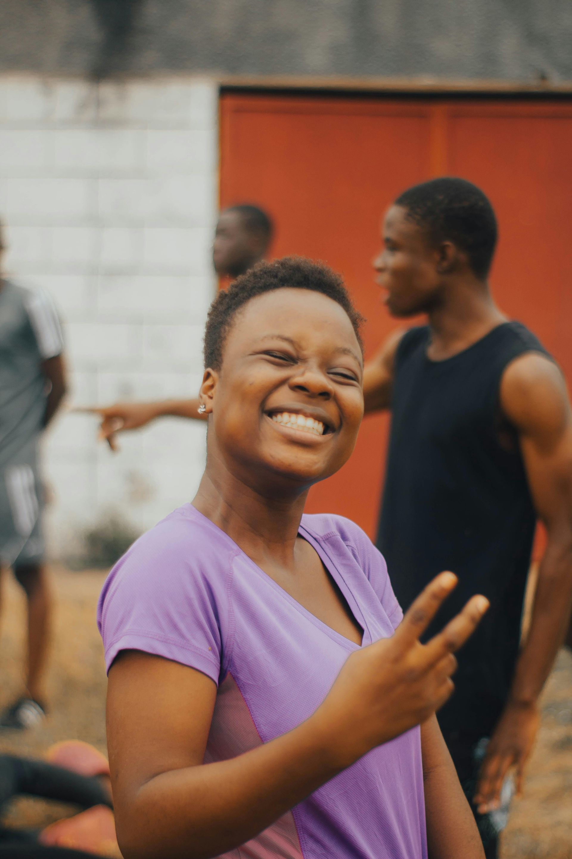 Smiling person in a purple shirt making a peace sign outdoors, with people in the background