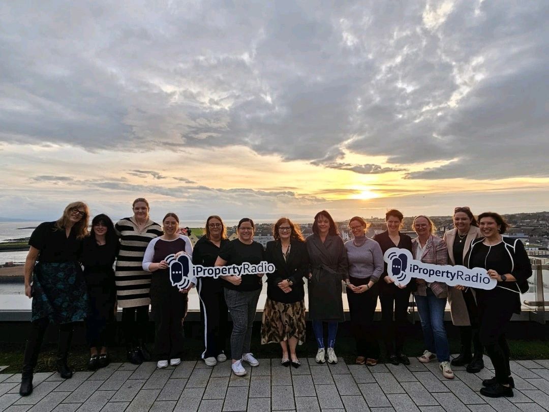 Group of people standing on a boardwalk at sunset, holding a banner with a logo and text.