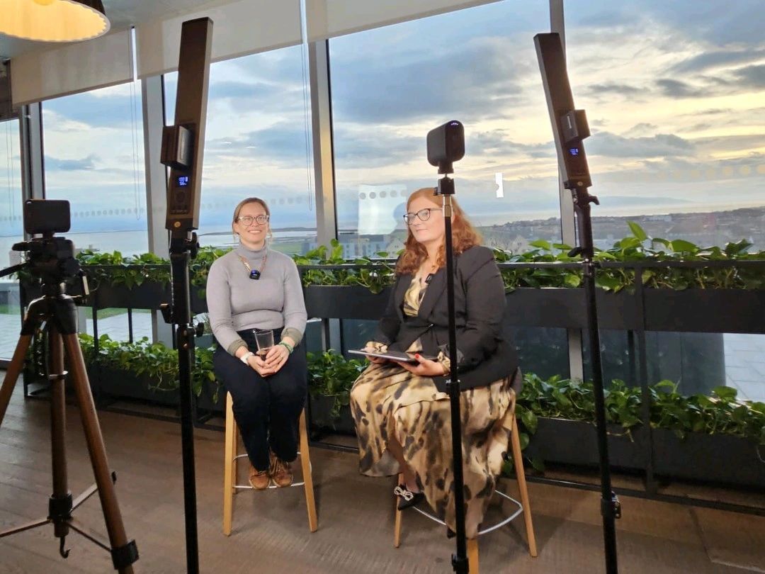 Two people seated on stools during an interview, with cameras on tripods and city view behind them.