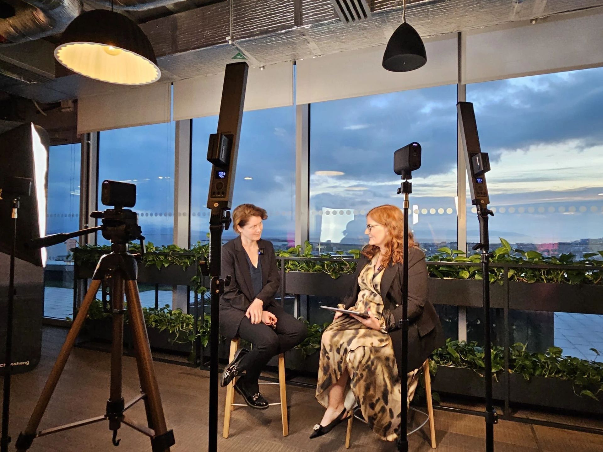Two women seated in a studio with tripods, lights, and a city skyline at sunset.