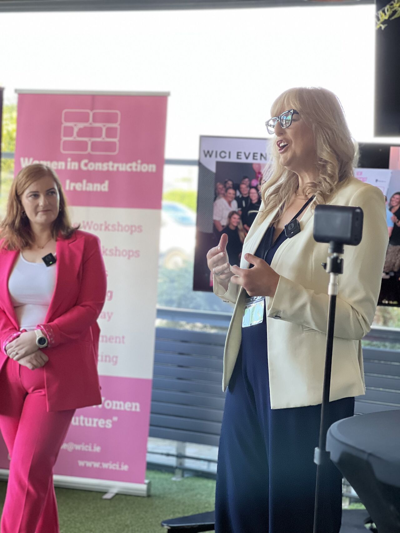 Two women speaking at a Women in Construction event, with pink banners in the background.