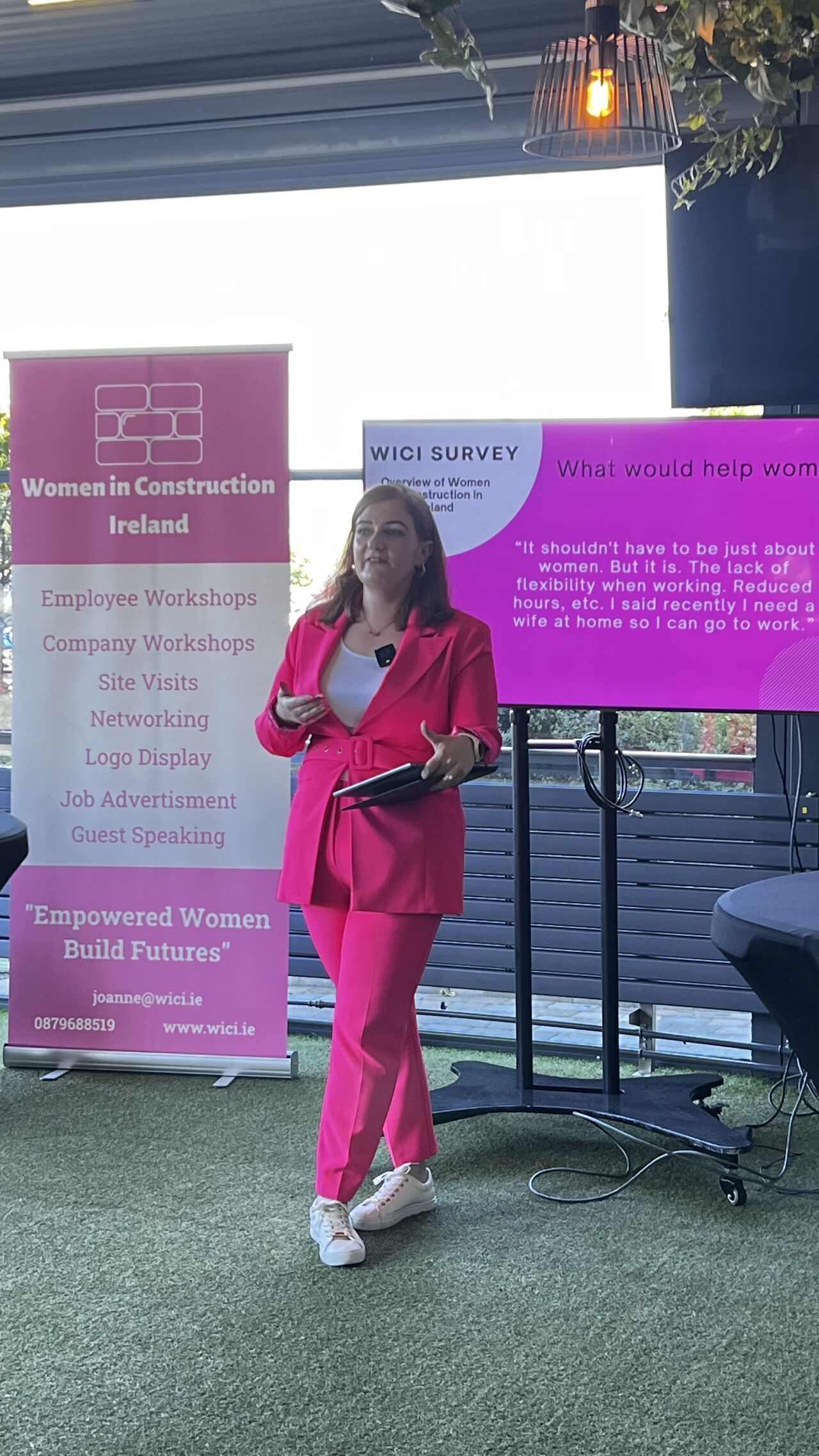 Woman in bright pink standing with microphone beside event banners and a presentation screen