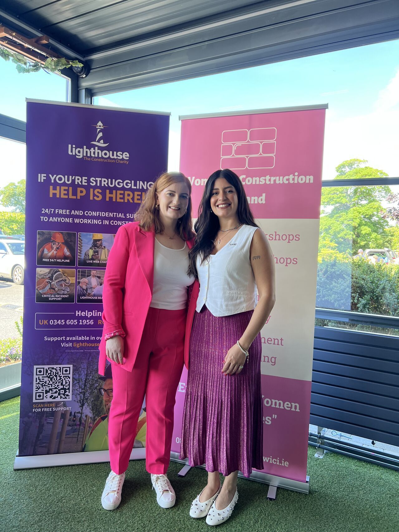 Two women standing at a promotional banner in pink outfits, smiling outdoors on a patio.