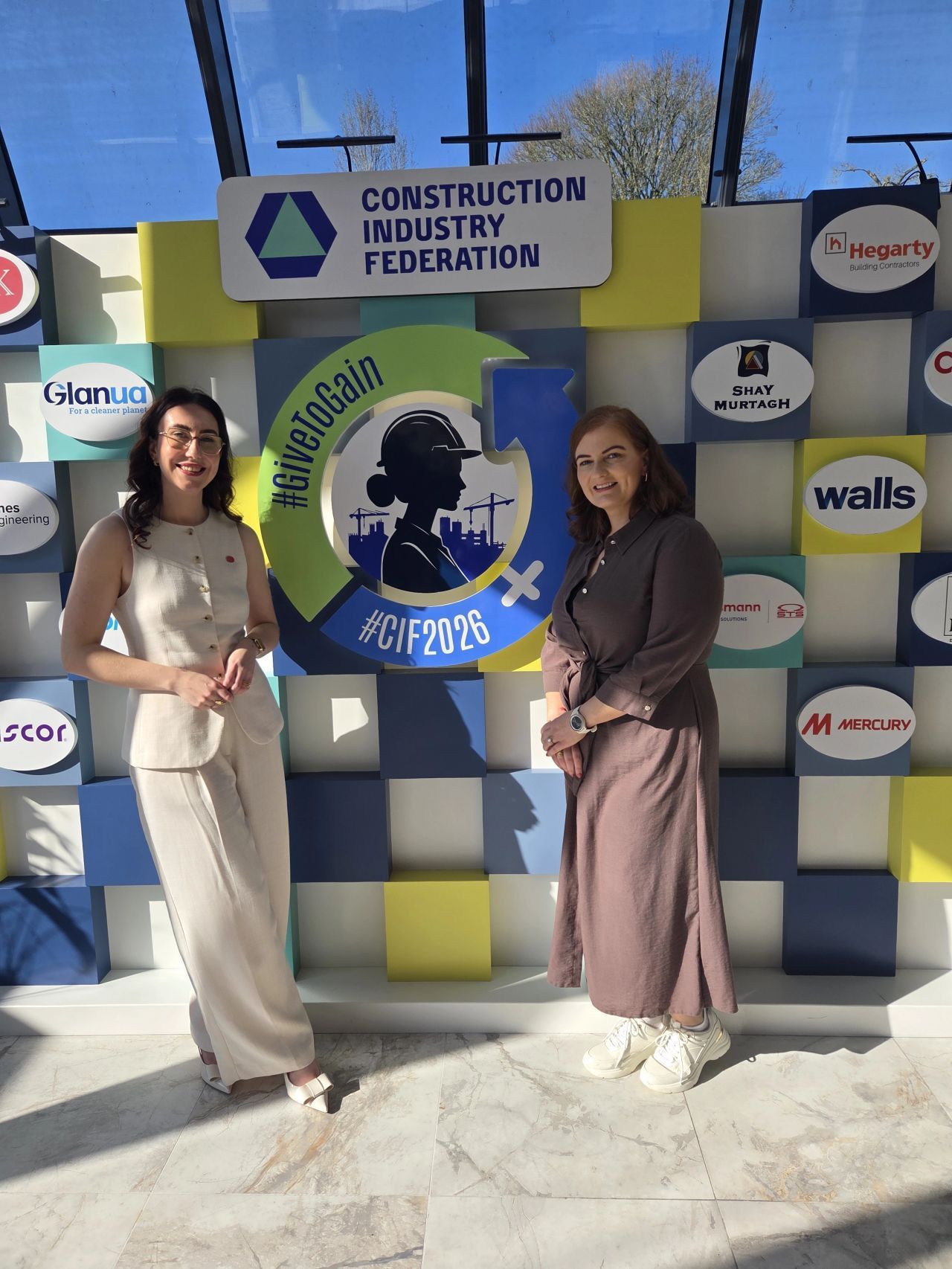 Two women posing at a Construction Industry Federation event backdrop with logos and blue-yellow panels