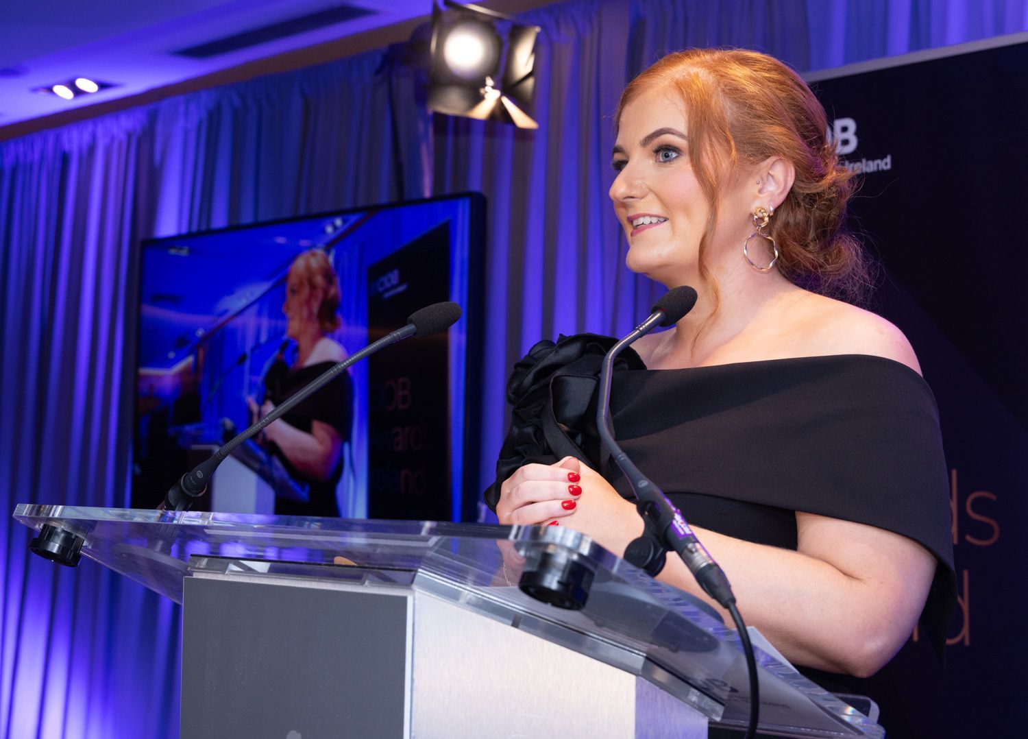 Woman speaking at a podium with a microphone, smiling under purple stage lights.