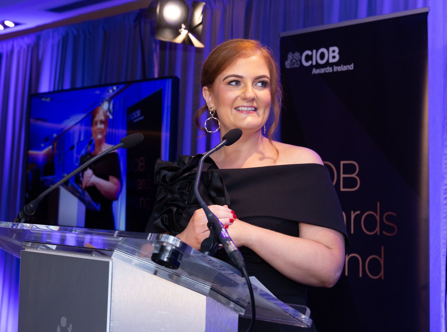 Woman speaking at a podium onstage at a CIOB awards event, with blue lighting and a large screen behind her.