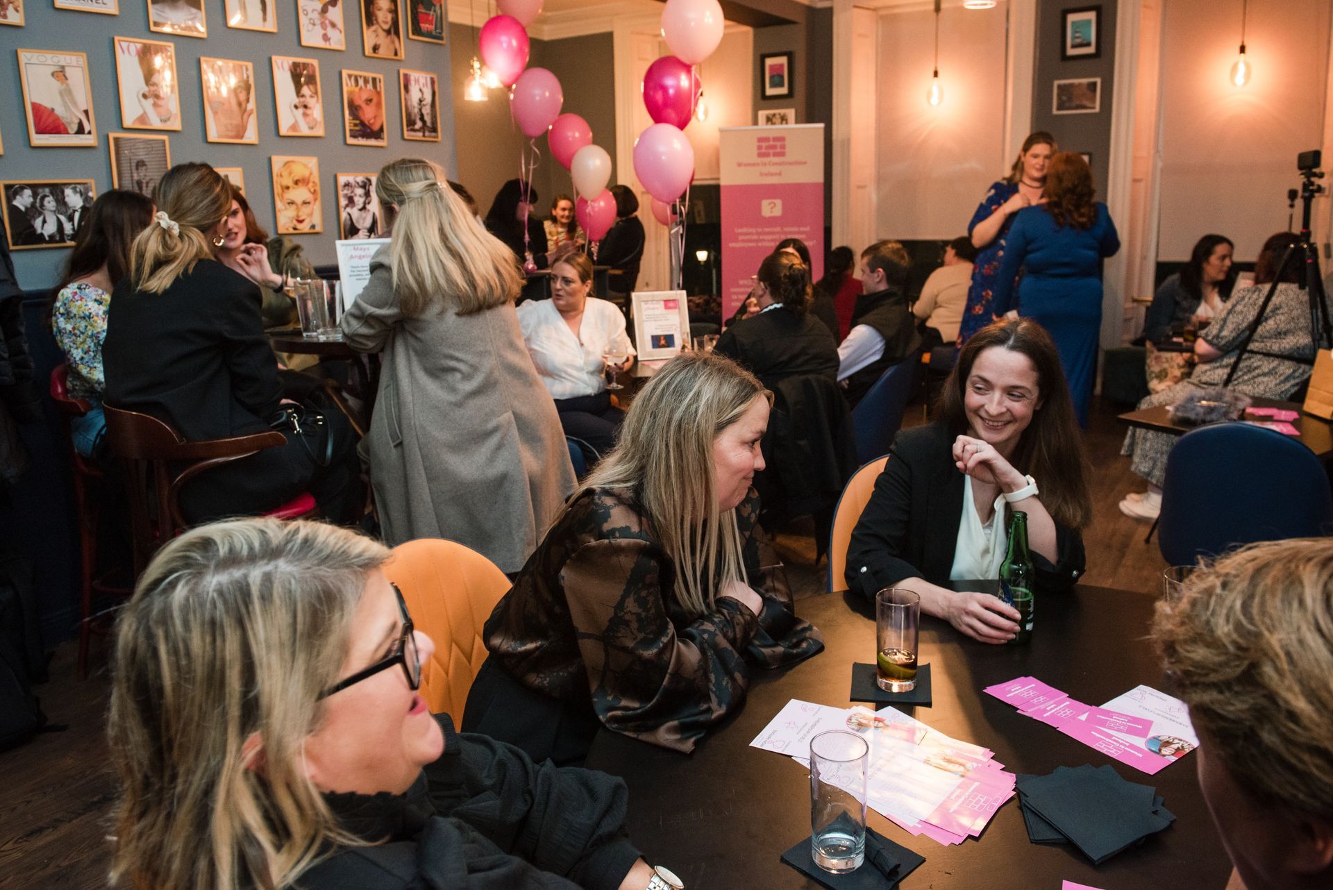 People chatting at a pink-themed event with balloons, framed photos, and tables with refreshments.