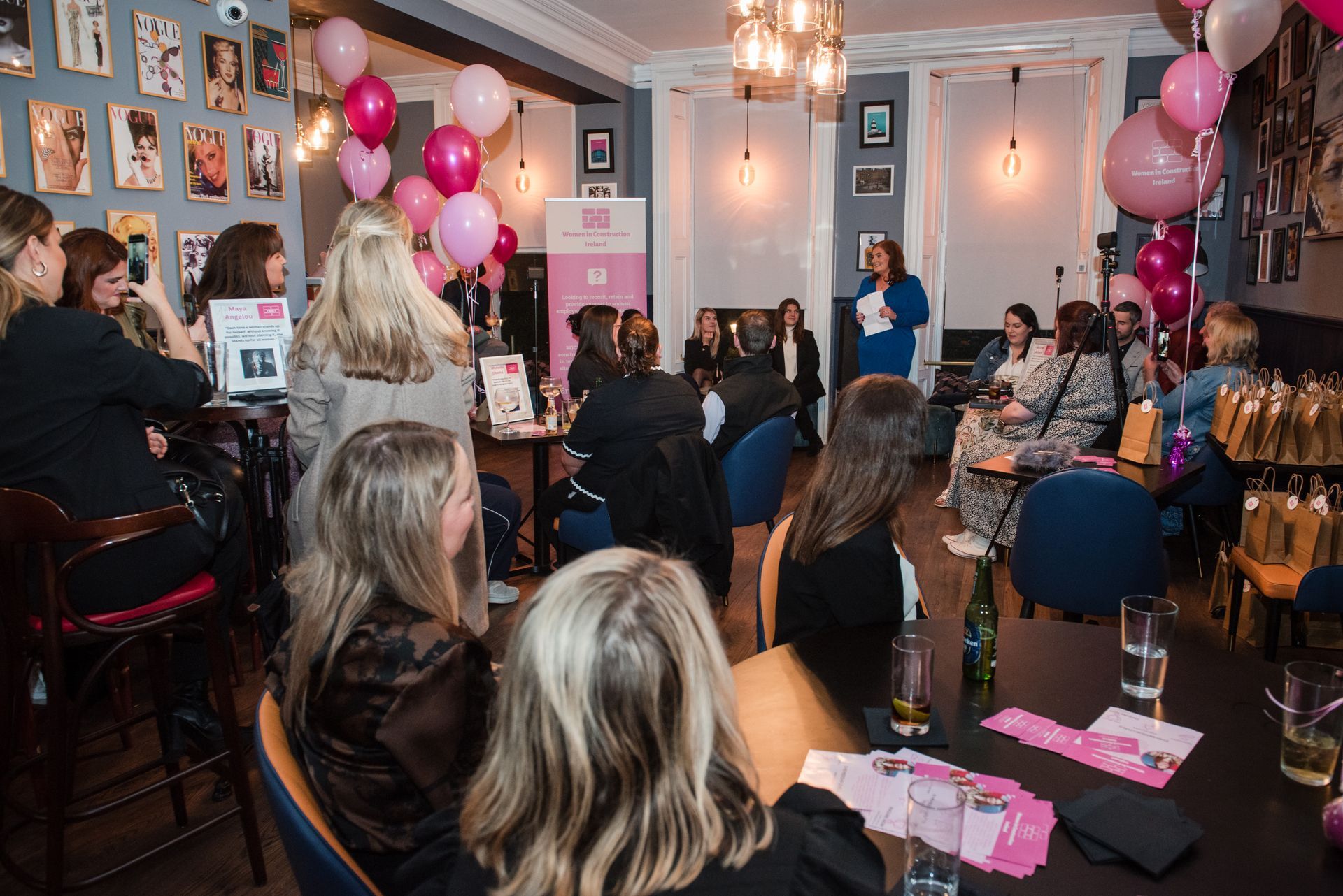 Crowded indoor event with pink balloons, people seated and standing, and a speaker at the front