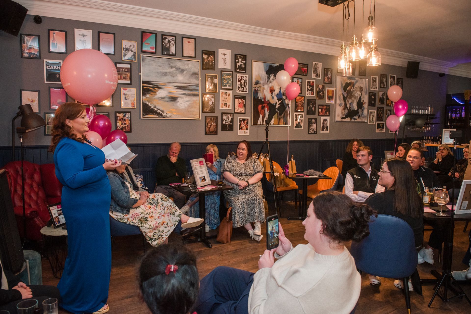 Birthday party in a cozy cafe, with guests seated and a woman holding pink balloons at the front.
