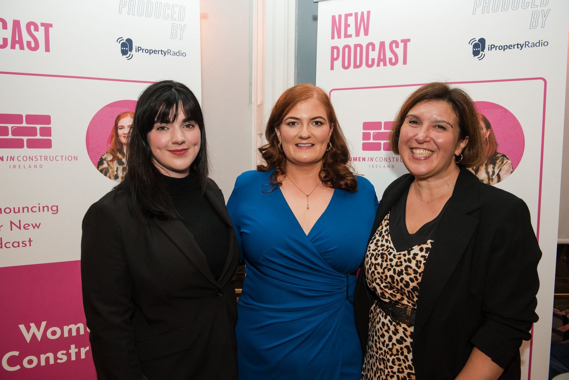Three women posing at a podcast launch event with pink and white branded backdrop.