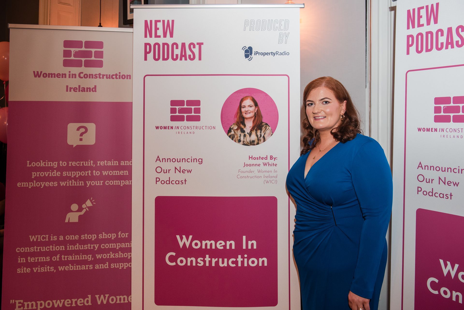 Woman standing beside pink “New Podcast” and “Women in Construction” display banners.