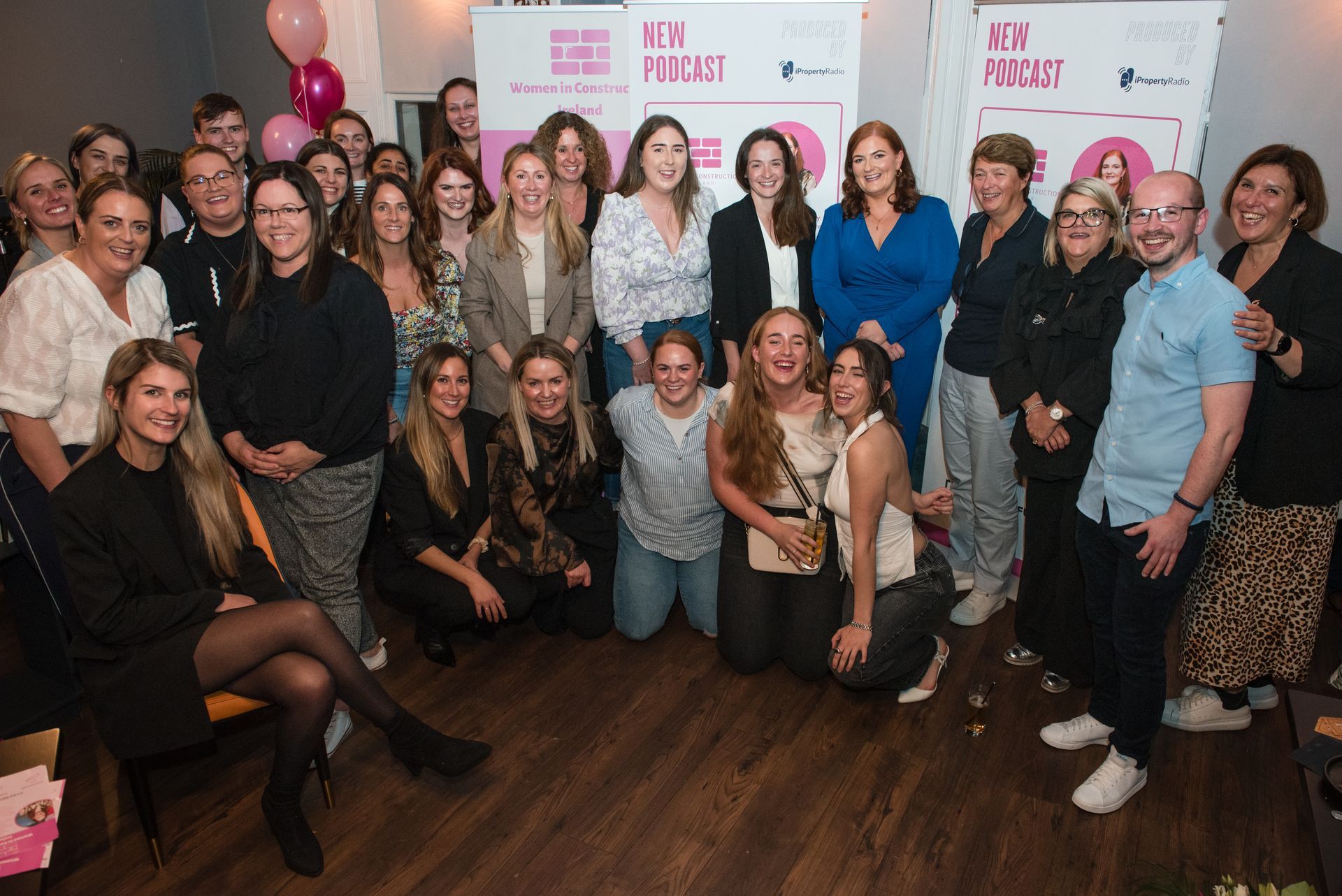Group of people posing at a pink-branded event backdrop, smiling indoors with balloons and drinks