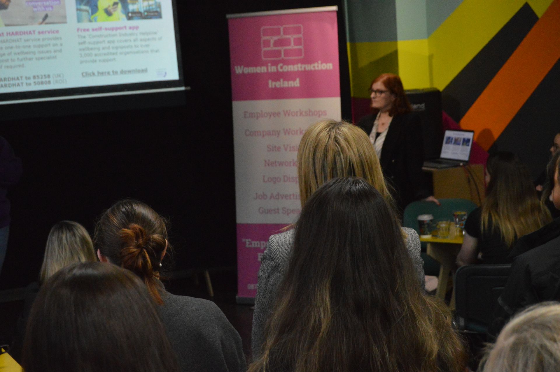 Audience watching a woman speak at a presentation beside a pink banner and projected slide.