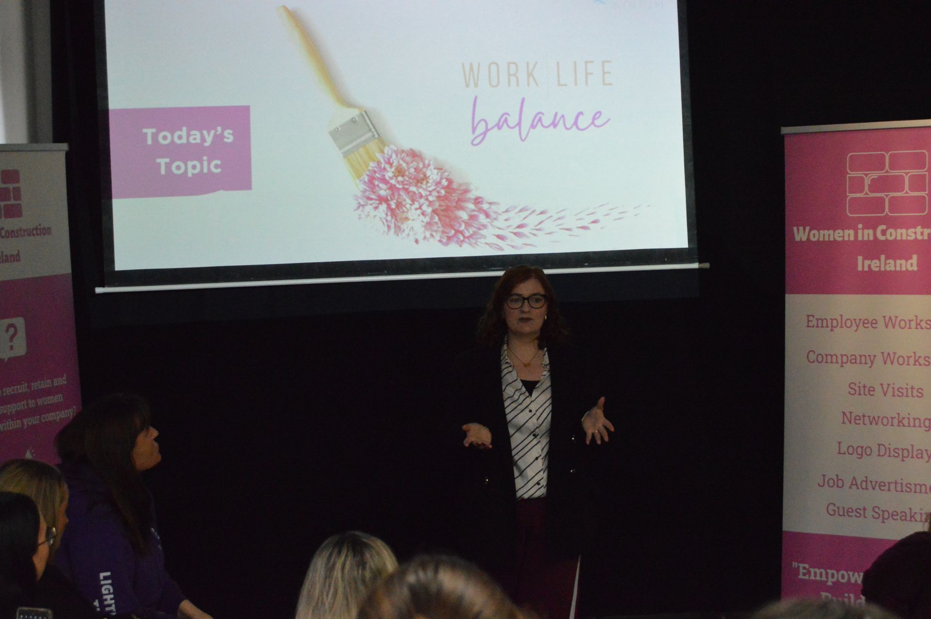 Presenter speaks on stage beside “Work Life Balance” slide and pink banners at a seminar