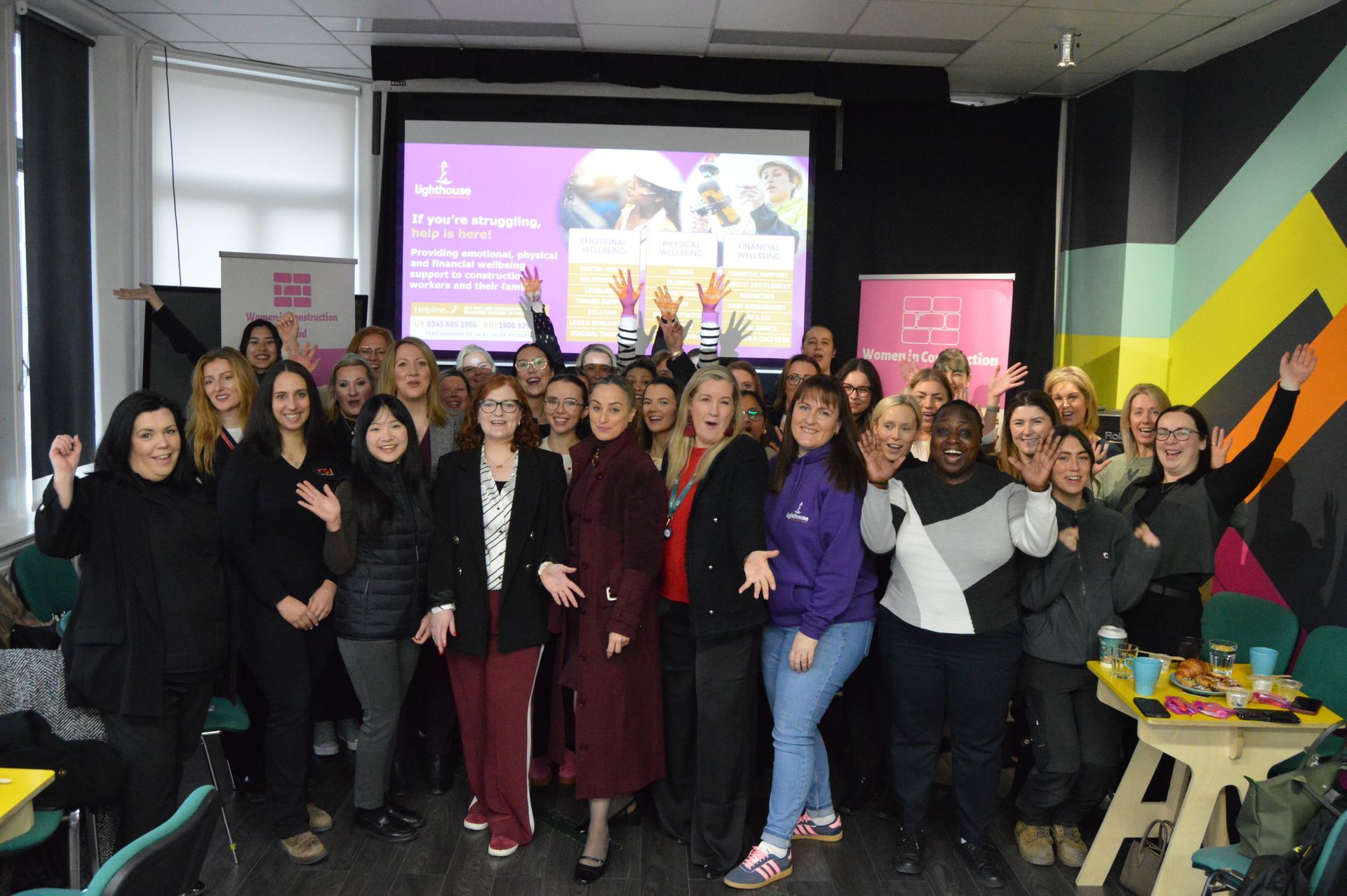 Group of people posing indoors at an event, with a presentation screen and colorful wall behind them.