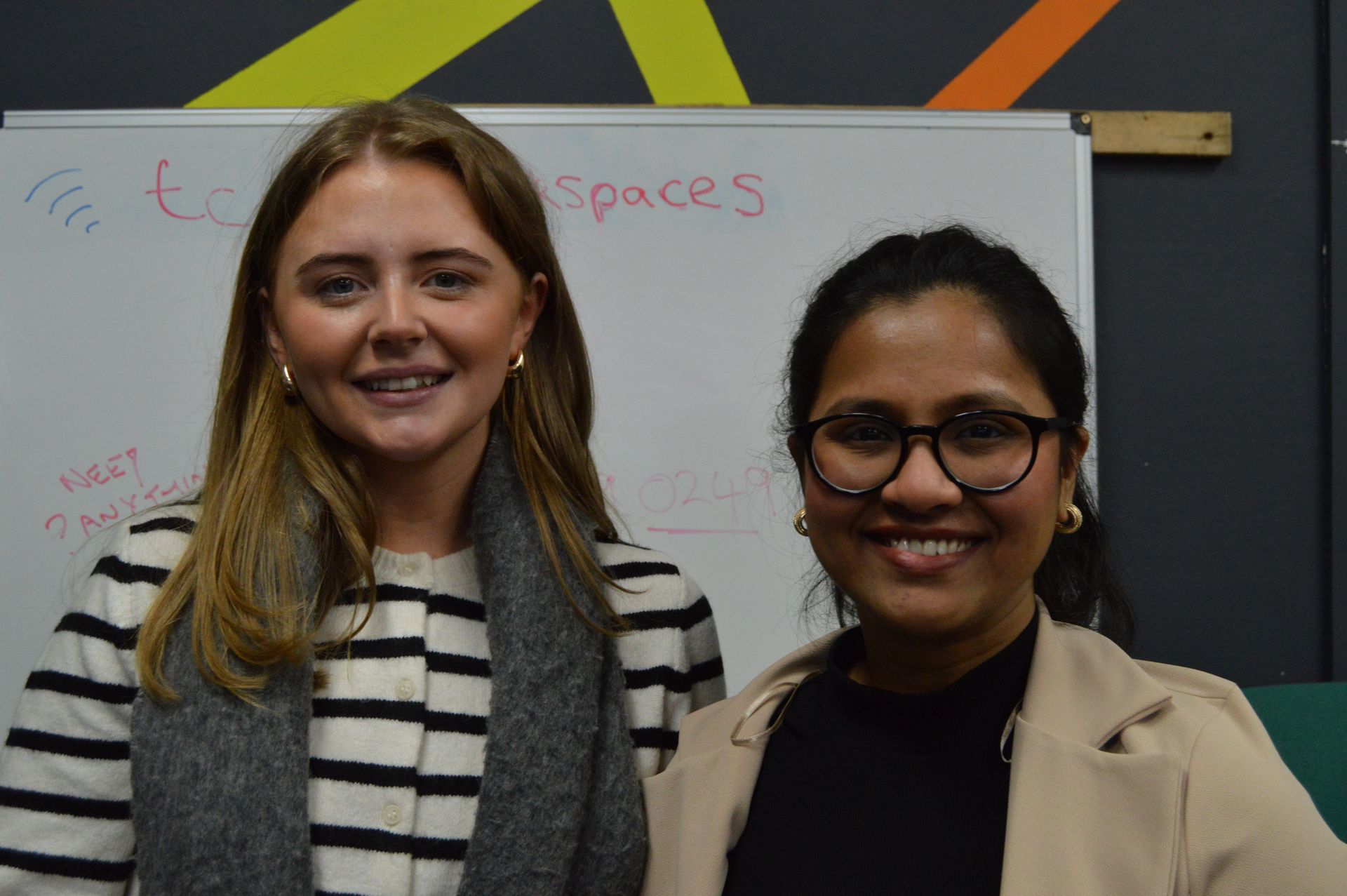 Two smiling people standing in front of a whiteboard in a classroom or office.