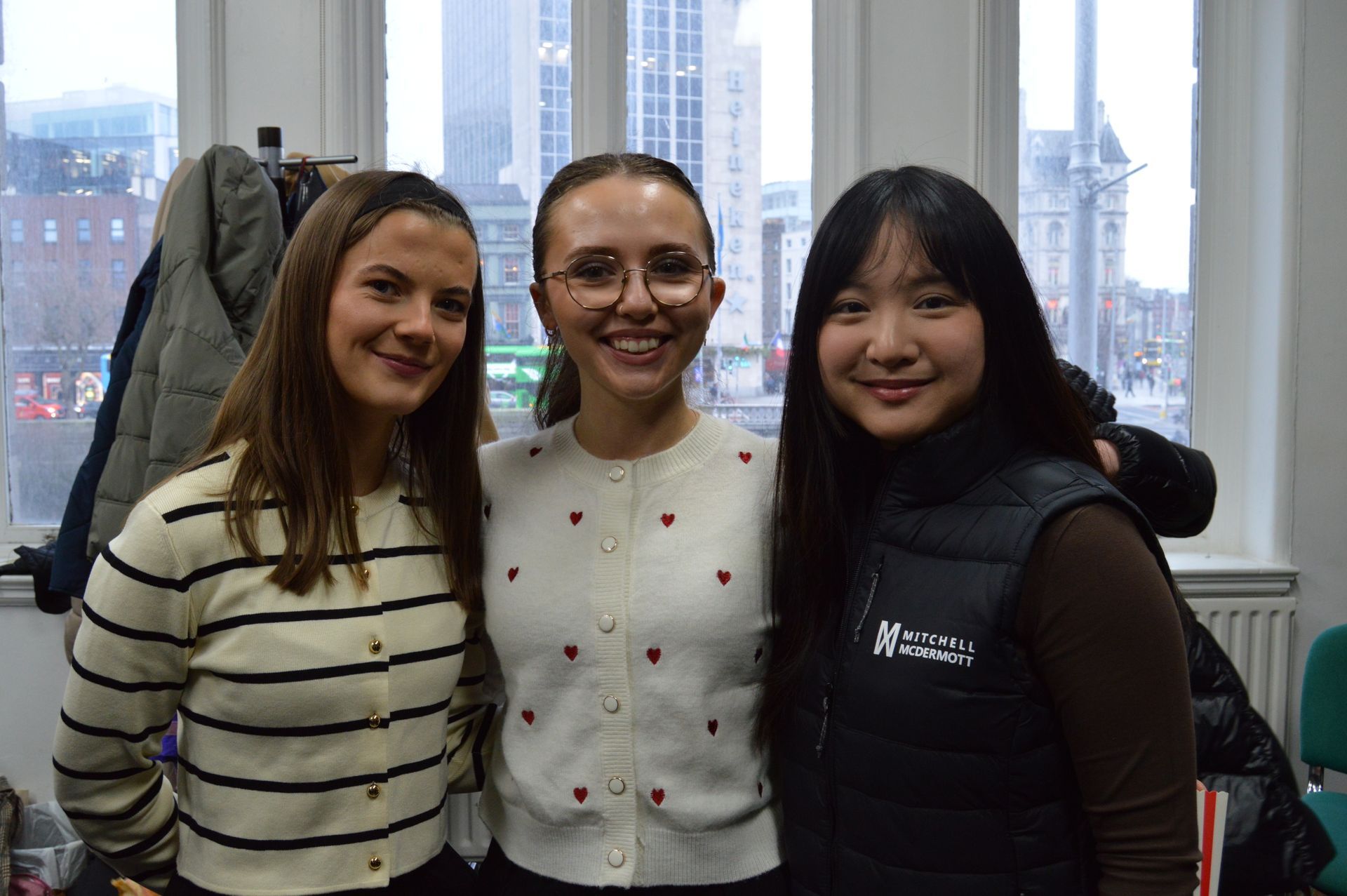 Three smiling friends standing indoors by a window with city buildings in the background