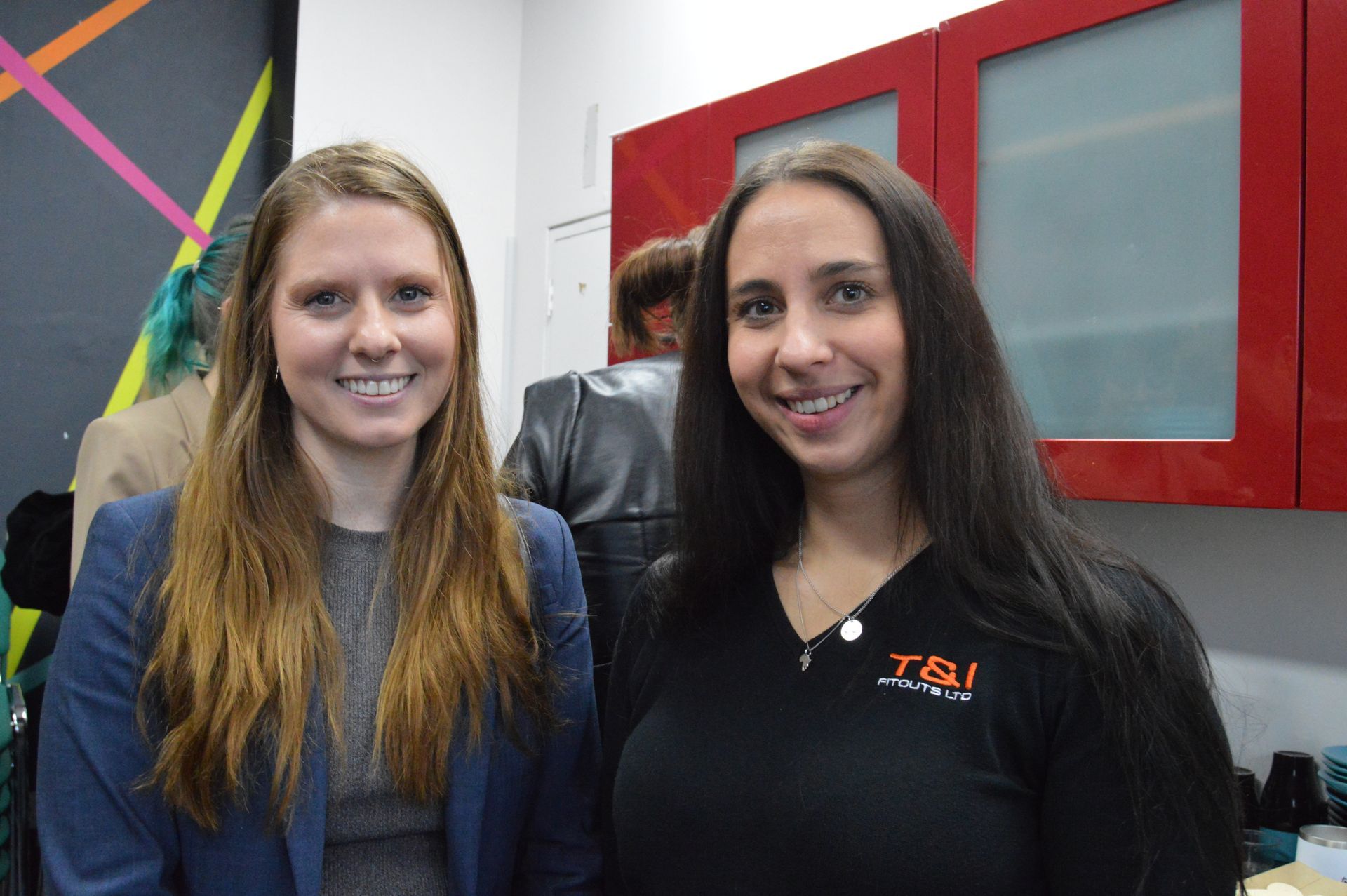 Two smiling women in an office, standing in front of a red-framed door.