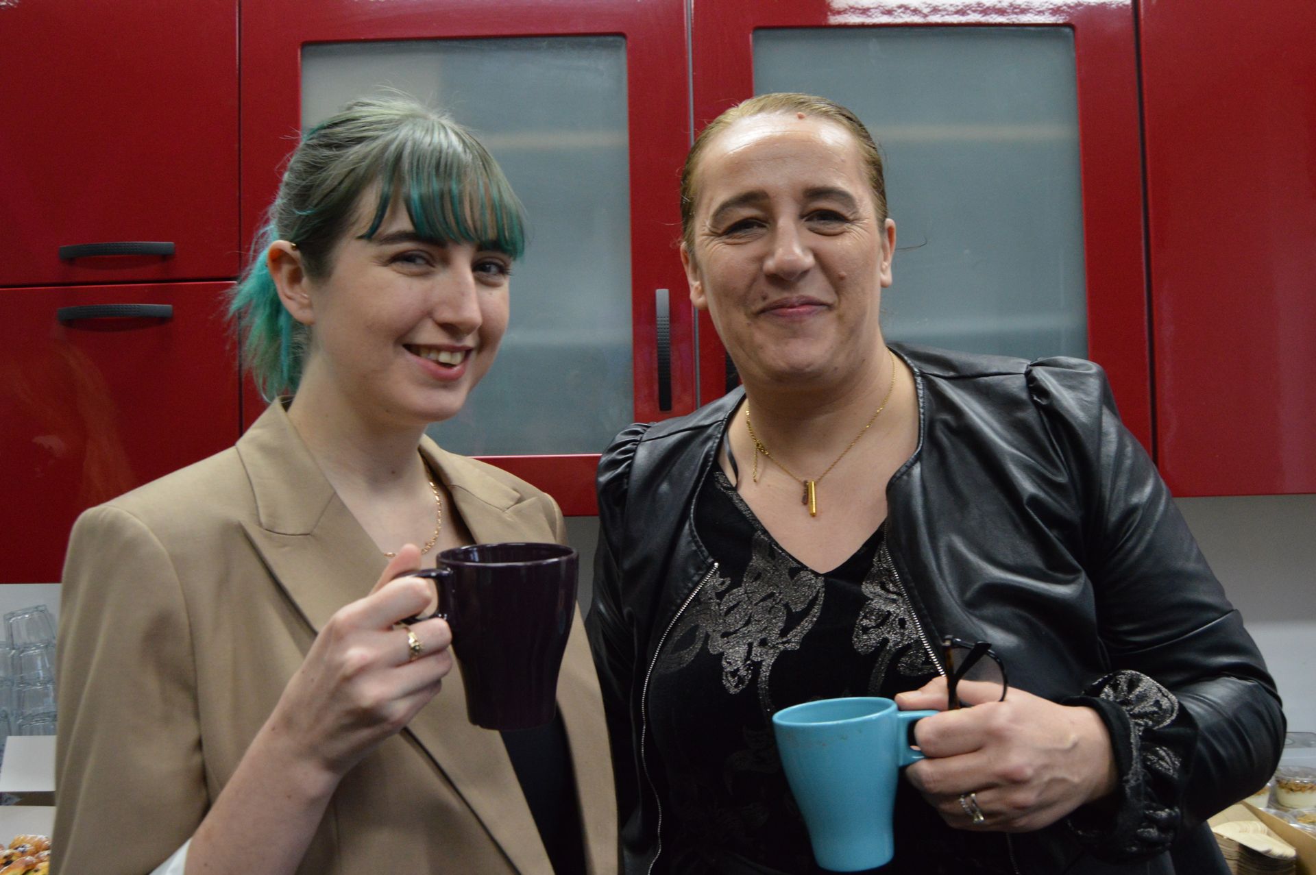 Two people smiling and holding mugs in front of a red wall and window