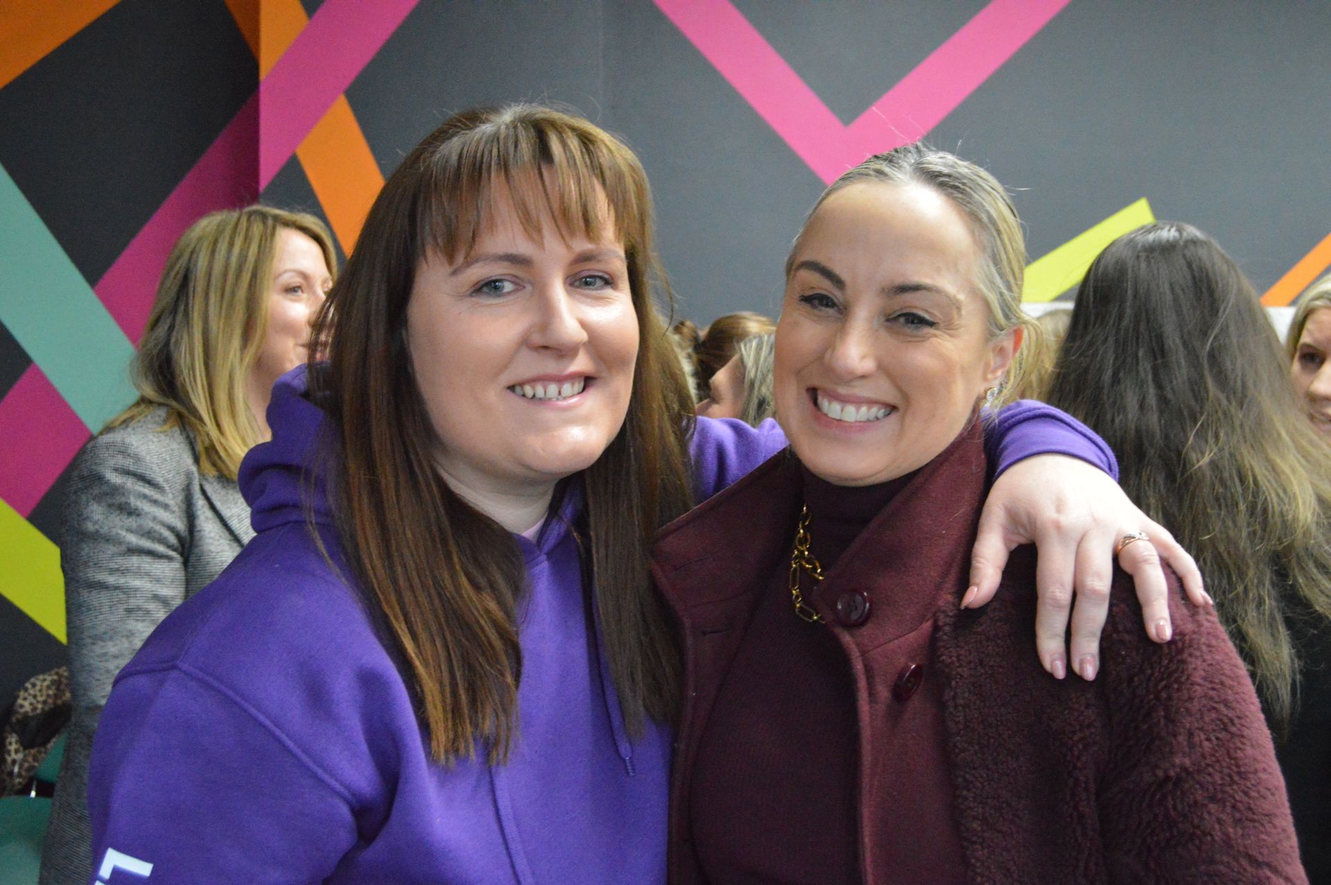 Two smiling people posing together indoors, one in a purple hoodie with an arm around the other.