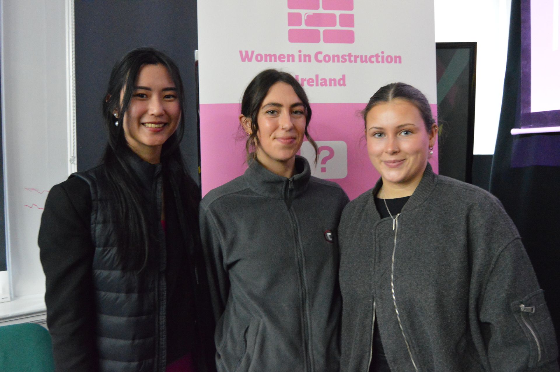 Three people standing indoors in front of a pink Women in Construction Ireland banner
