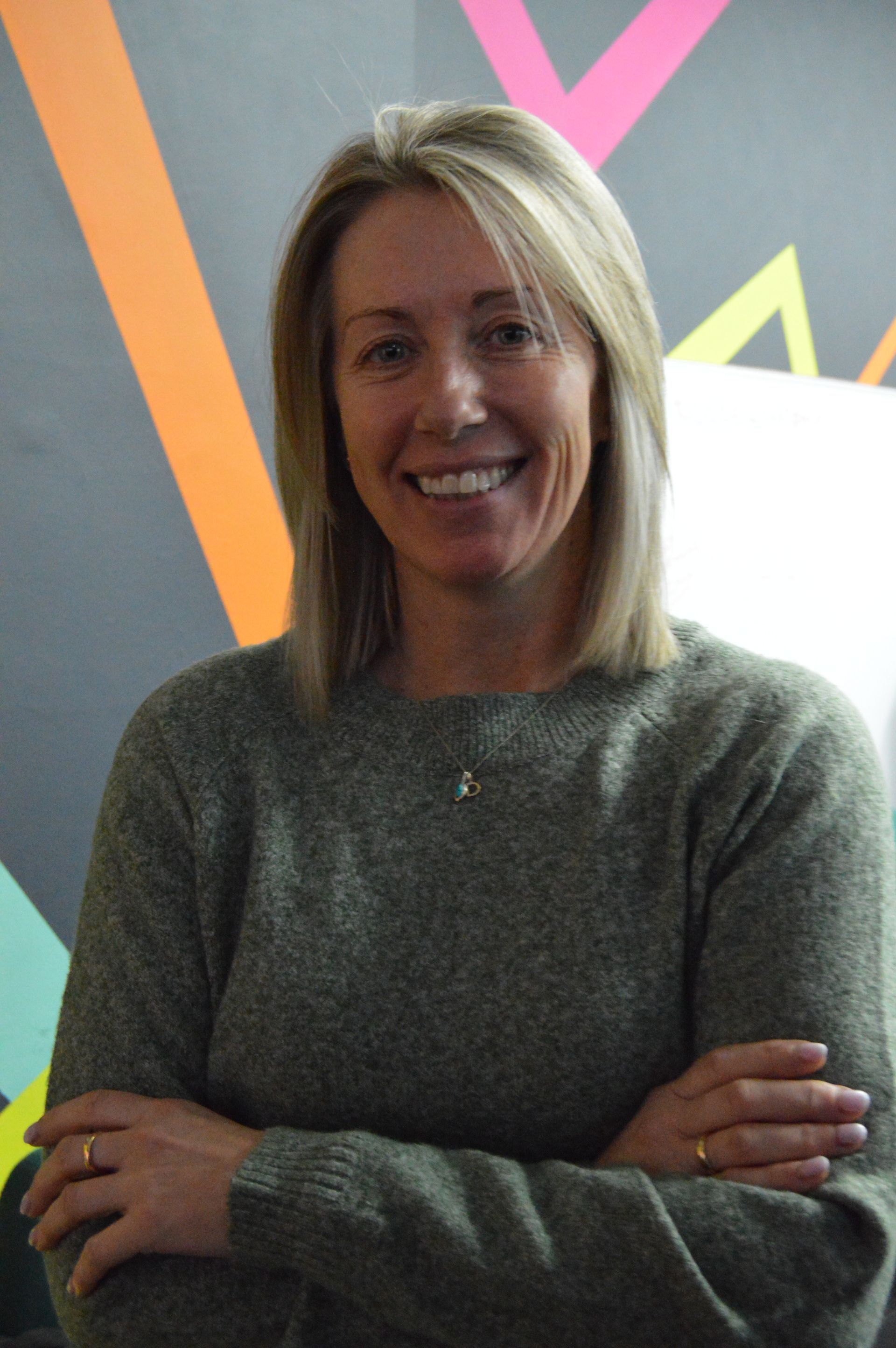 Woman smiling with arms crossed, wearing a gray sweater in front of a colorful wall