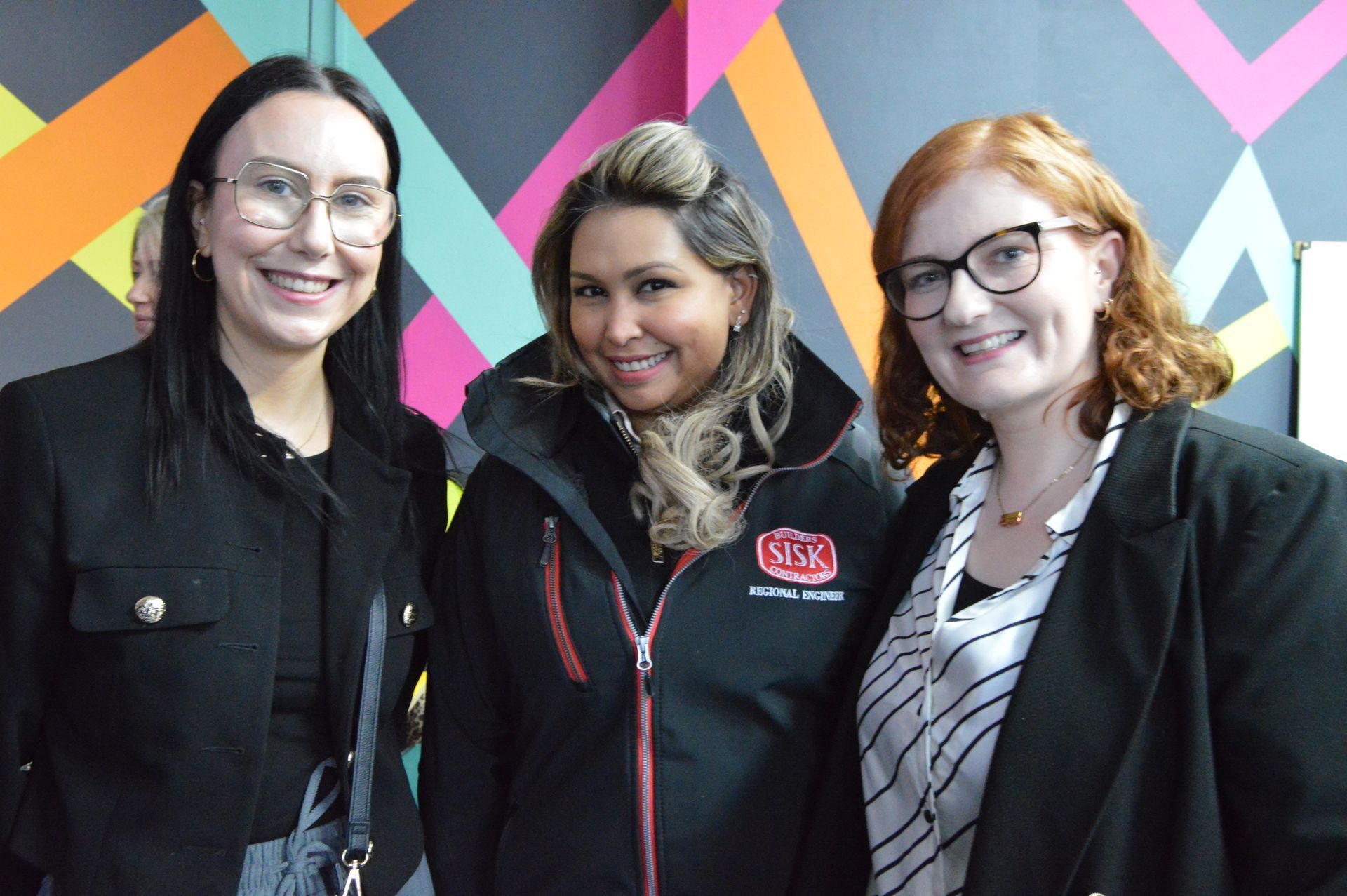 Three smiling people standing indoors in front of a colorful geometric wall