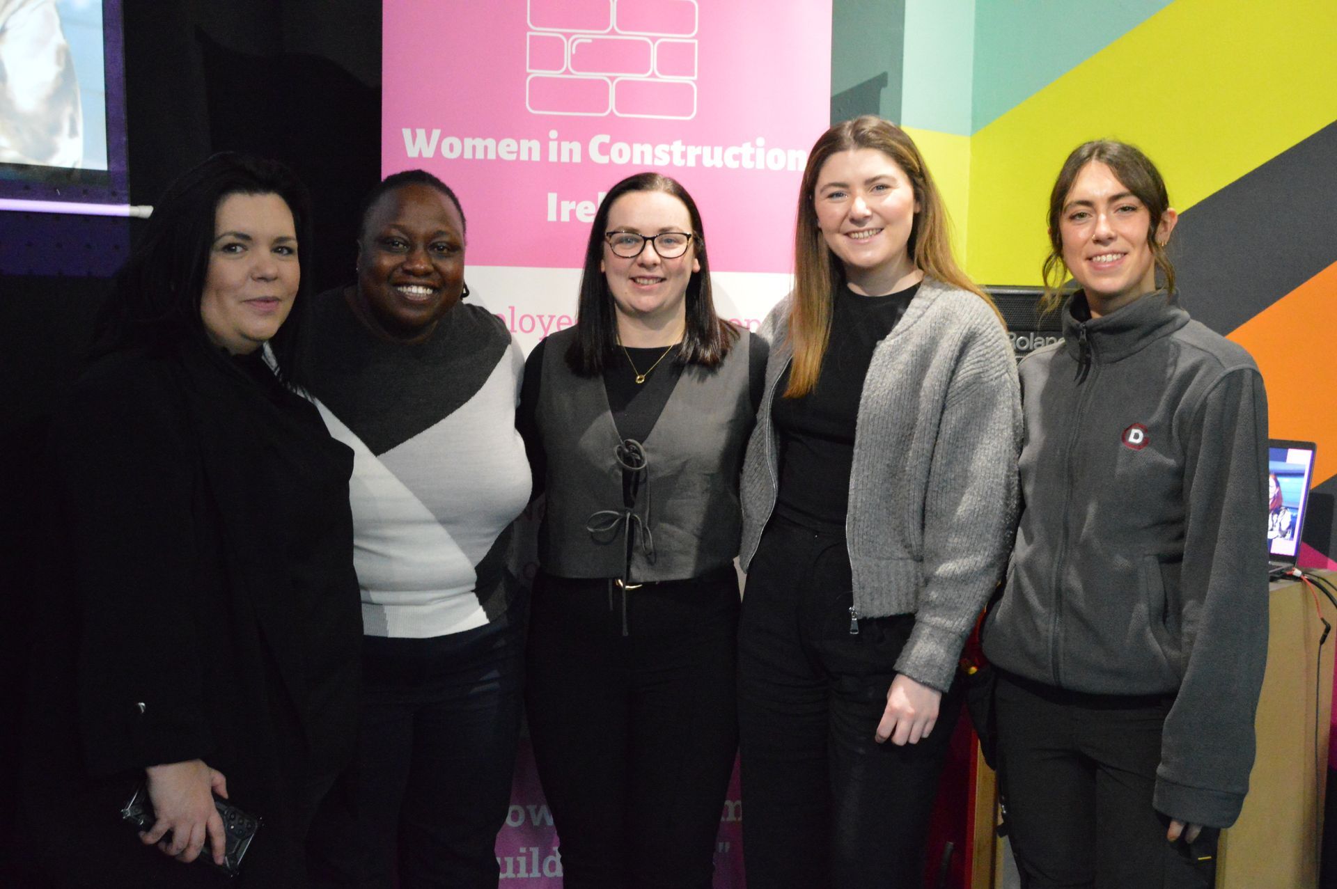 Five people standing together at a Women in Construction event banner indoors