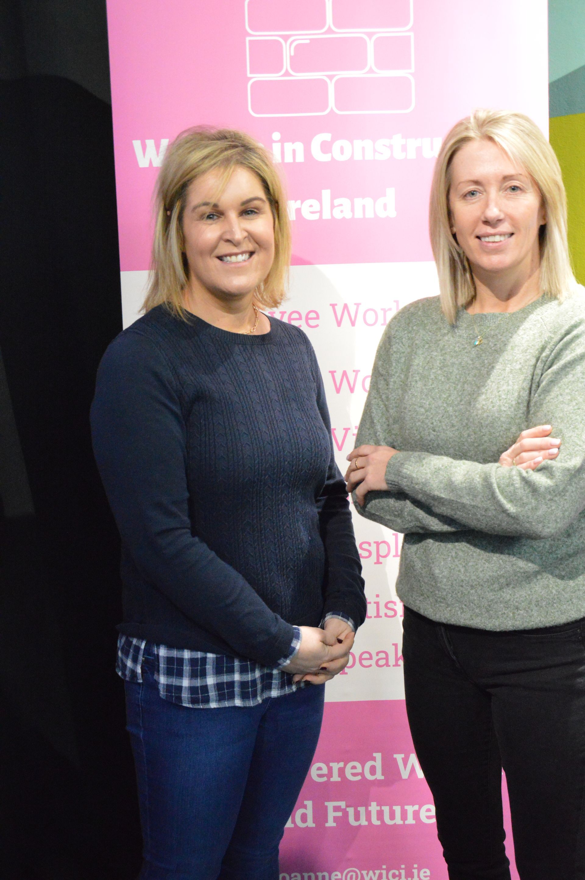 Two women standing in front of a pink banner, smiling with arms crossed.