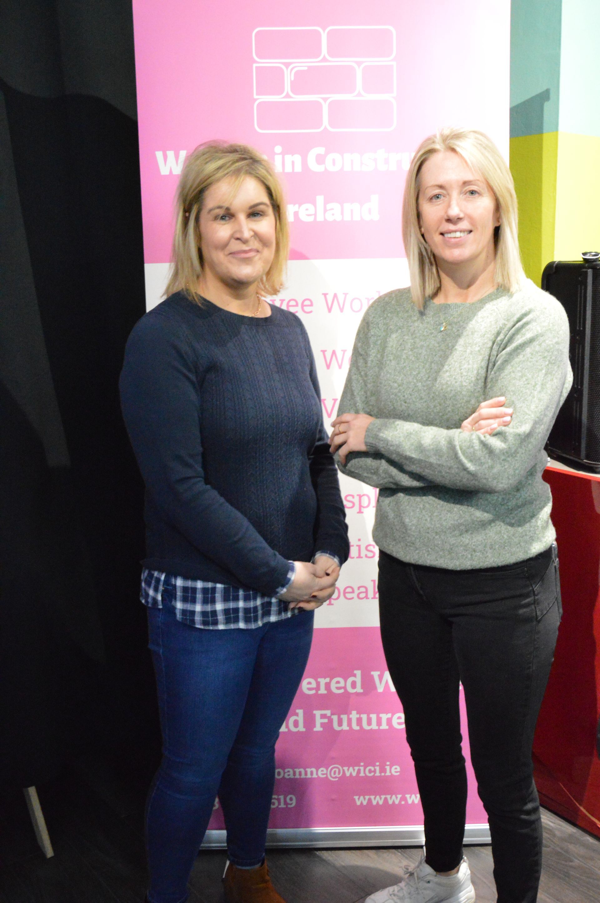 Two women standing beside a pink event banner, posing with arms crossed.