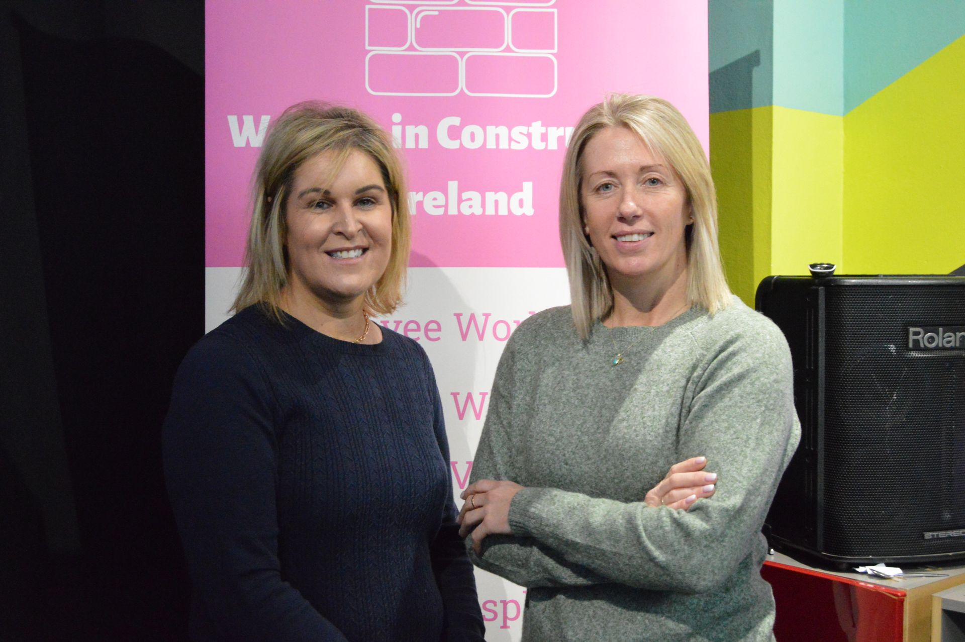 Two women stand in front of a pink event banner, arms crossed and smiling.