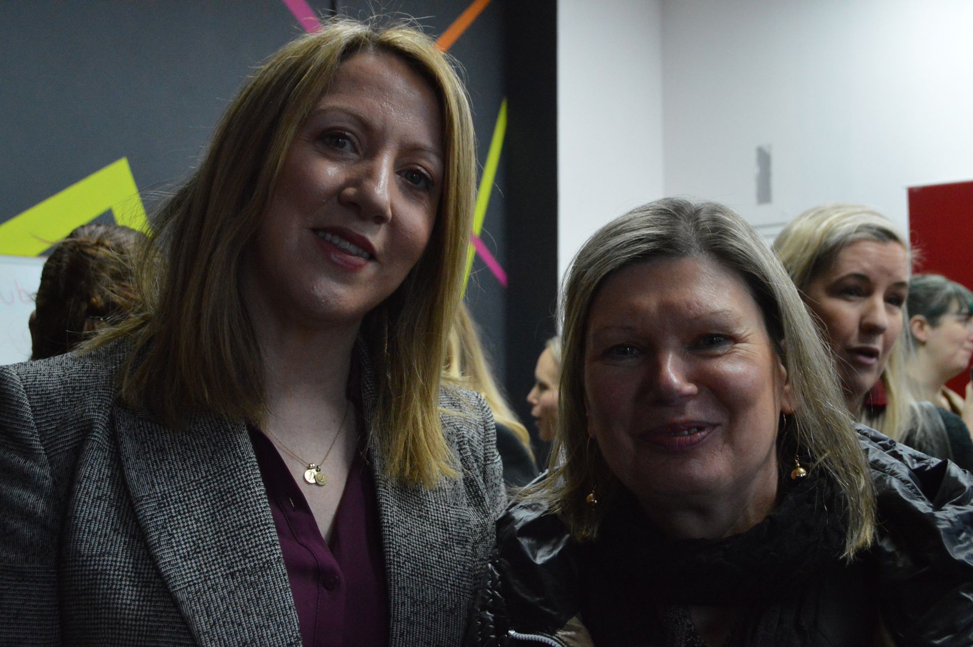 Two people smiling at an indoor event, with a dark wall and geometric accents in the background