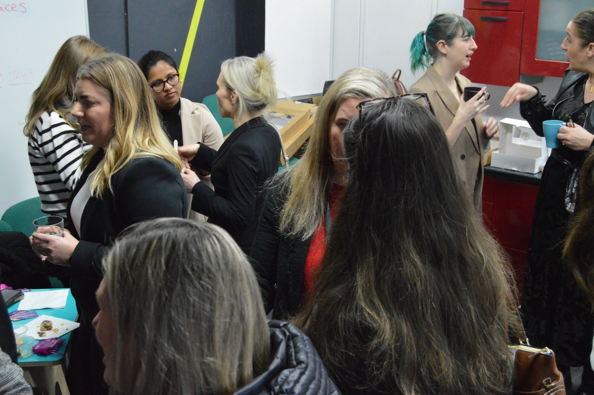Crowded indoor event with people chatting beside red and gray walls, one holding a tray of drinks