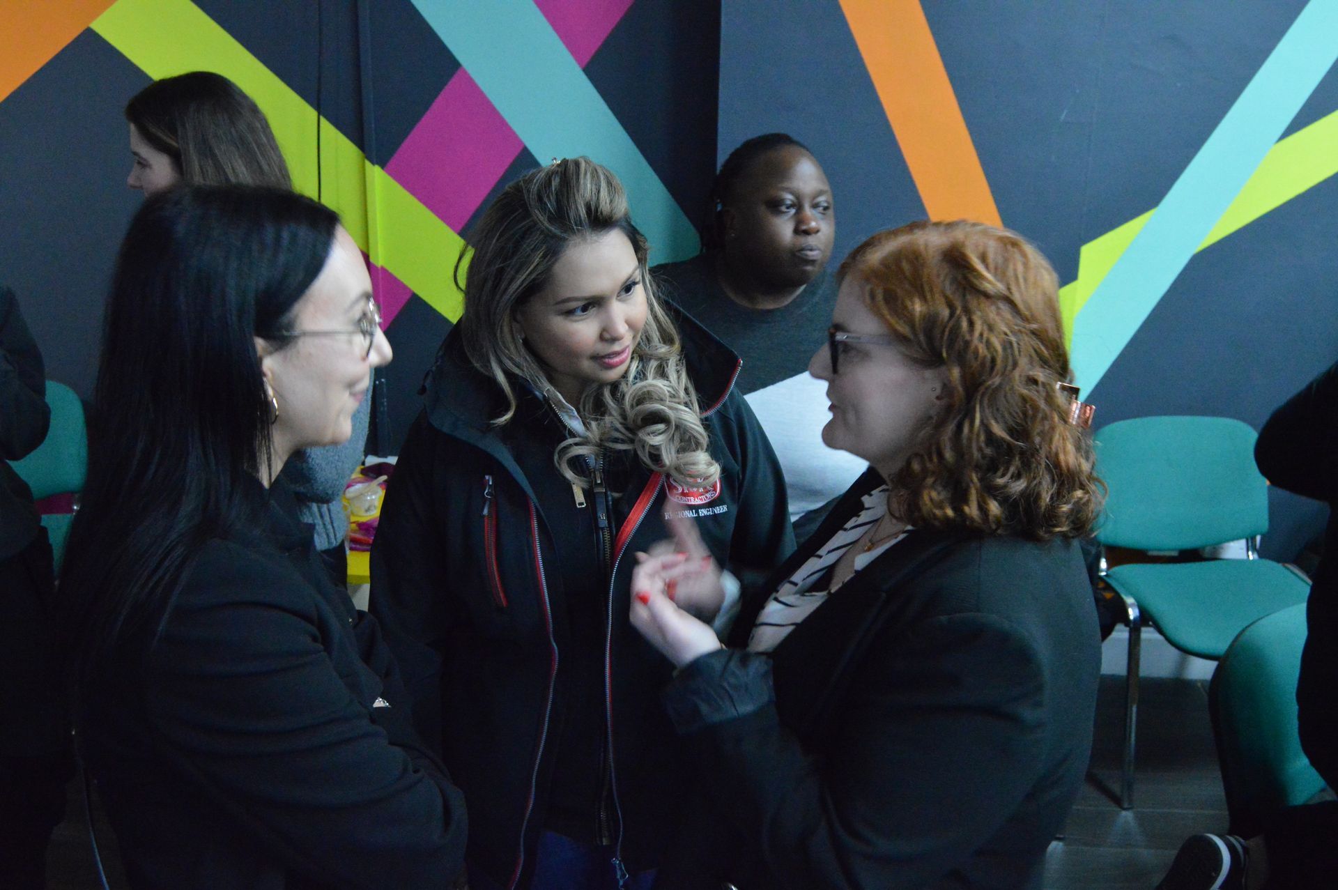 Four people talking in front of a colorful geometric mural indoors.