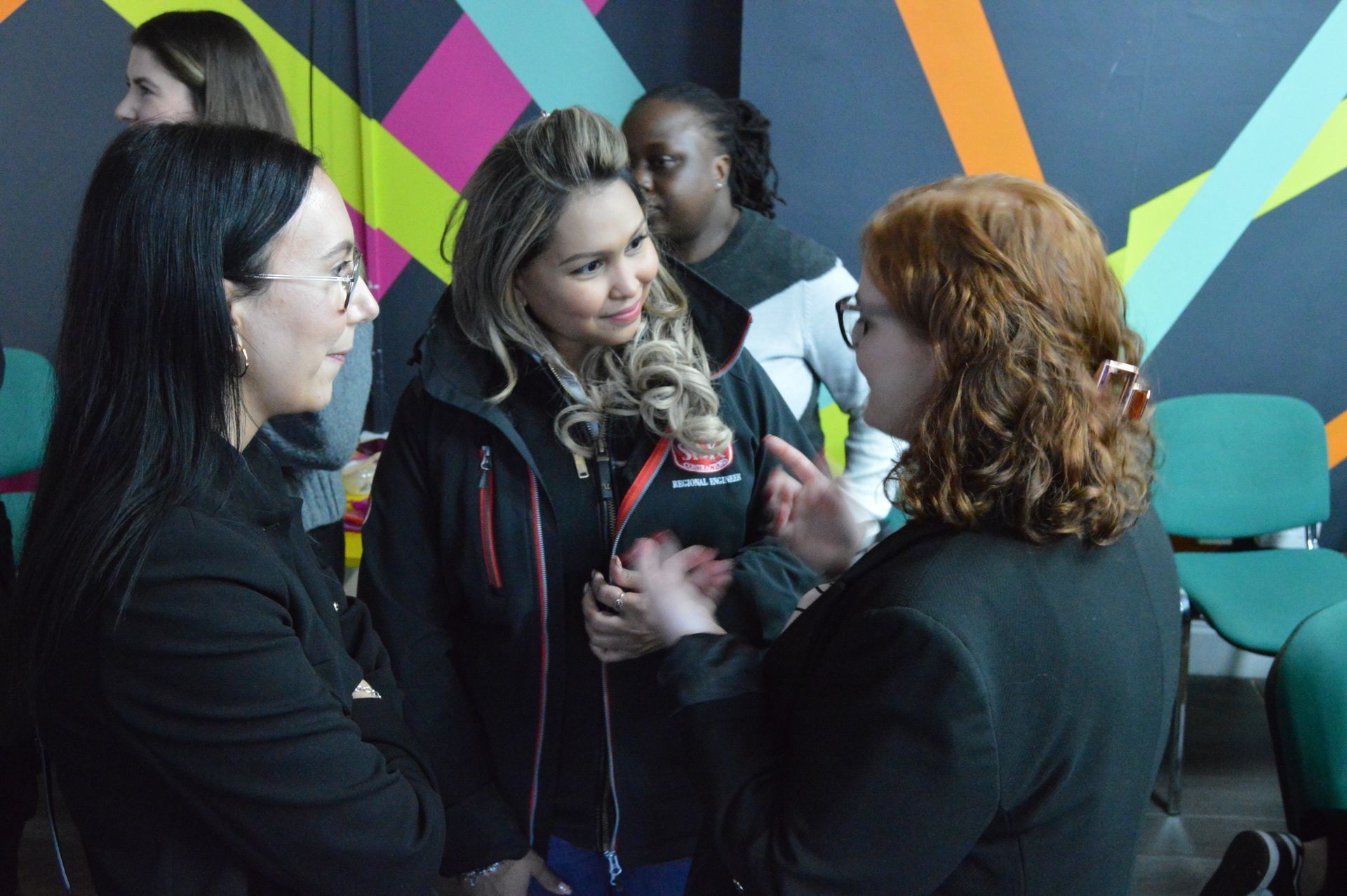 Four people chatting indoors in front of a colorful geometric wall and teal chairs.