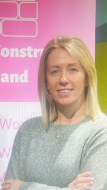 Blonde woman in a gray sweater stands with arms crossed in front of a pink event backdrop.