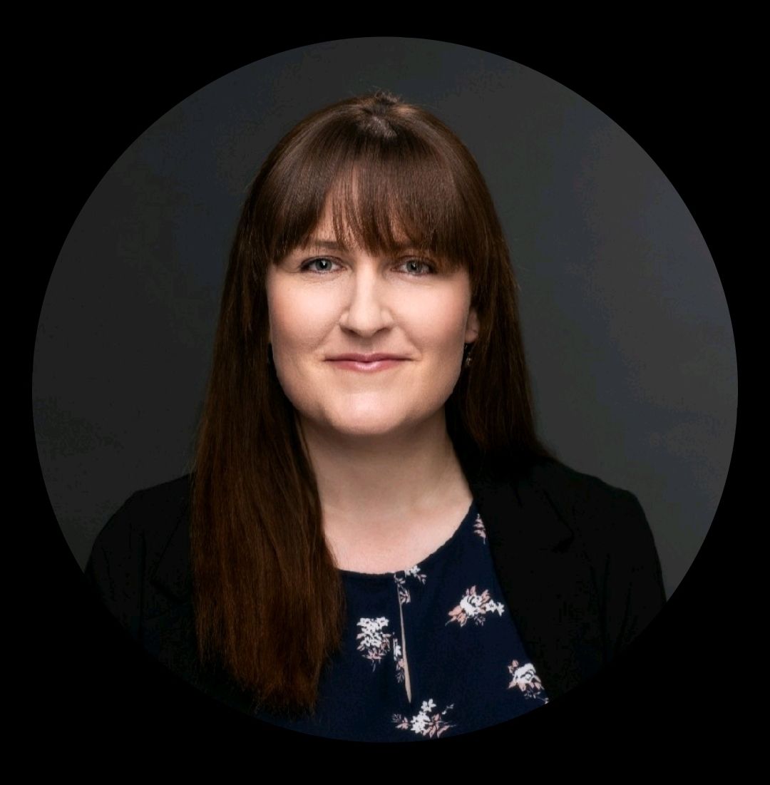 Studio portrait of a woman with long brown hair, wearing a black blazer and floral top, against a dark background