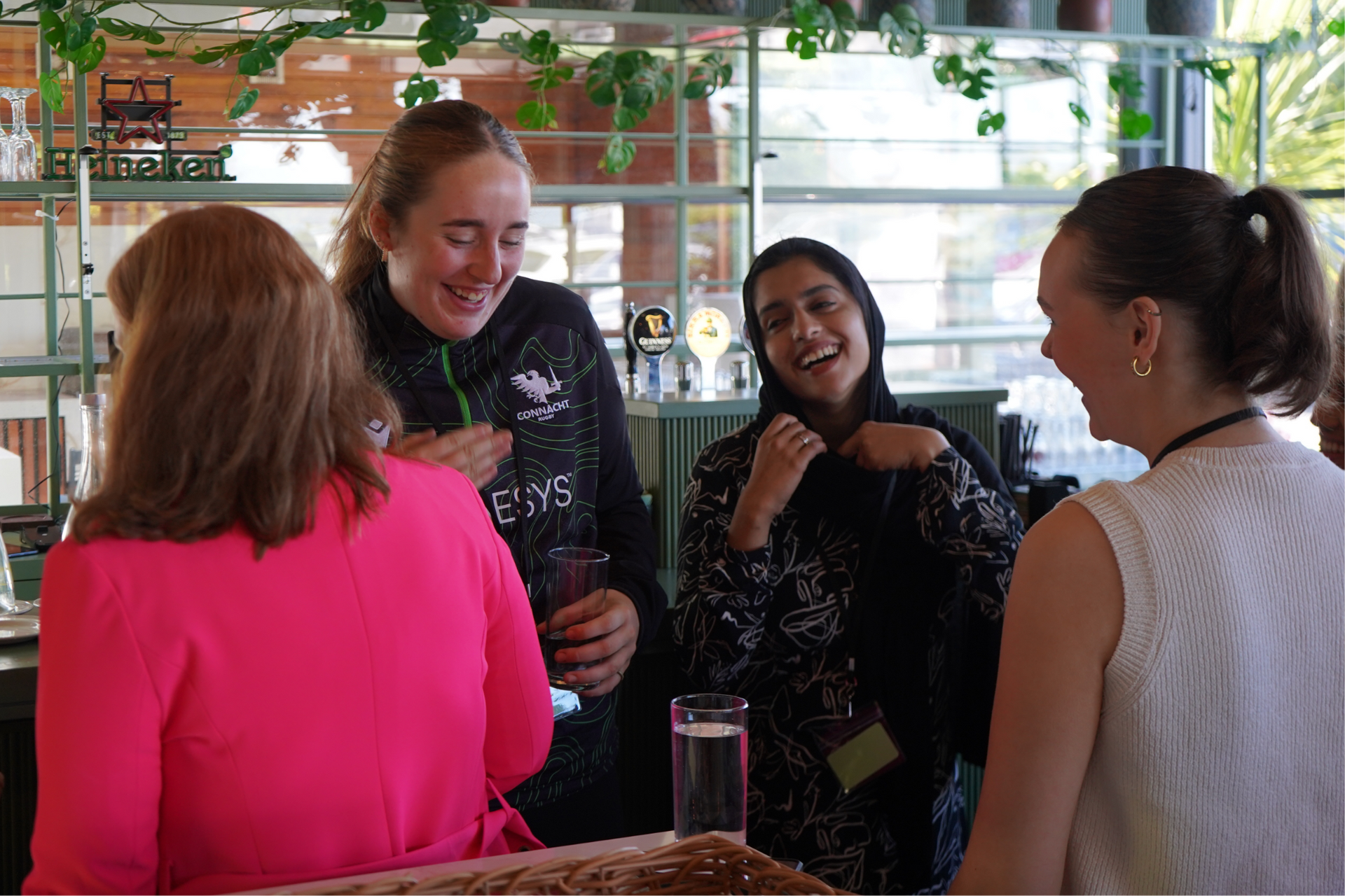 Four people chat and smile around a table in a bright café or bar with plants and shelves in the background.