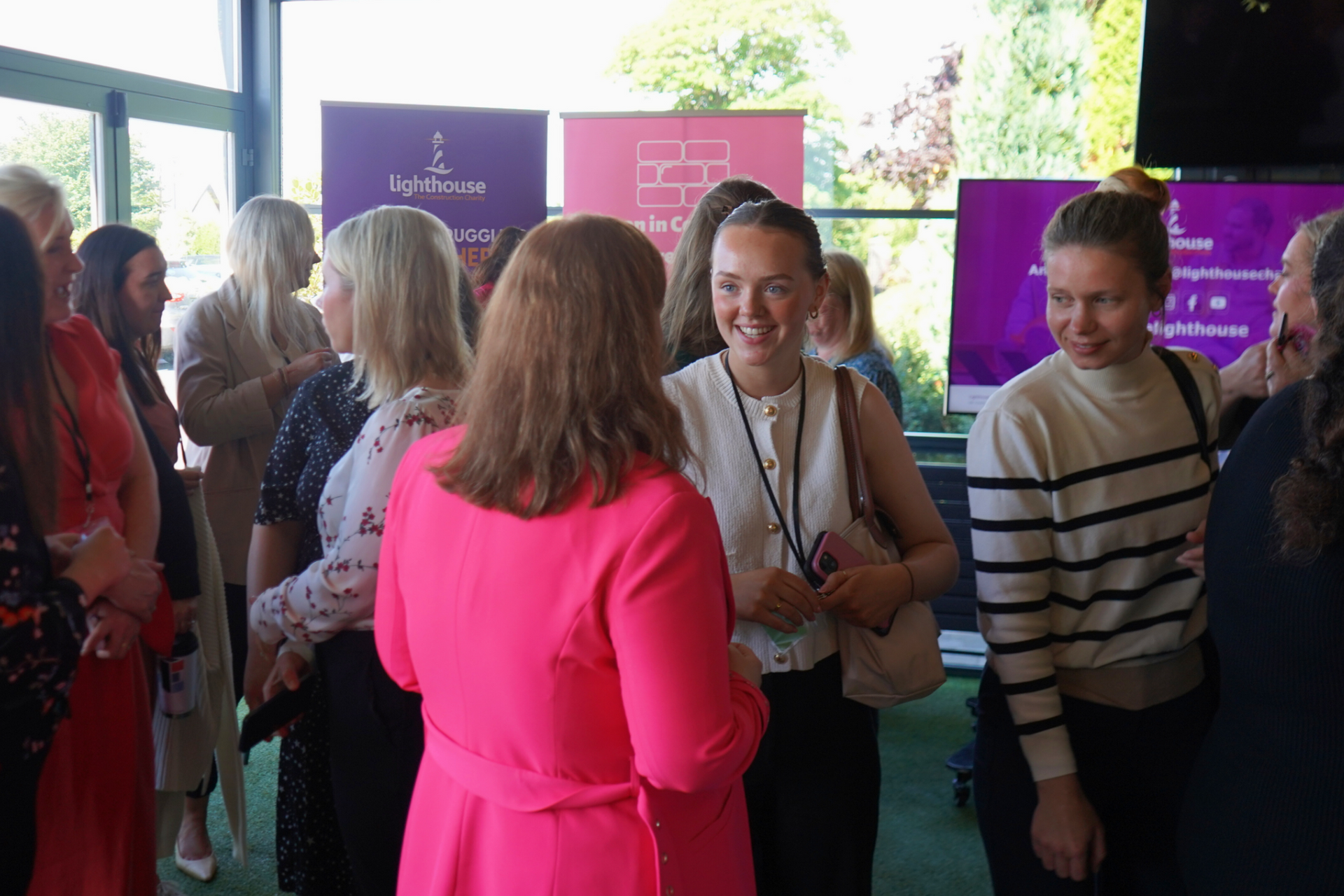 People chatting at an indoor event, with purple signs and a woman in a bright pink jacket in the foreground