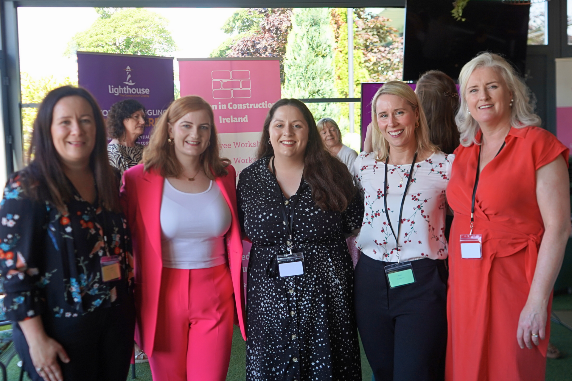 Five women standing together at an outdoor event, smiling in front of informational banners.