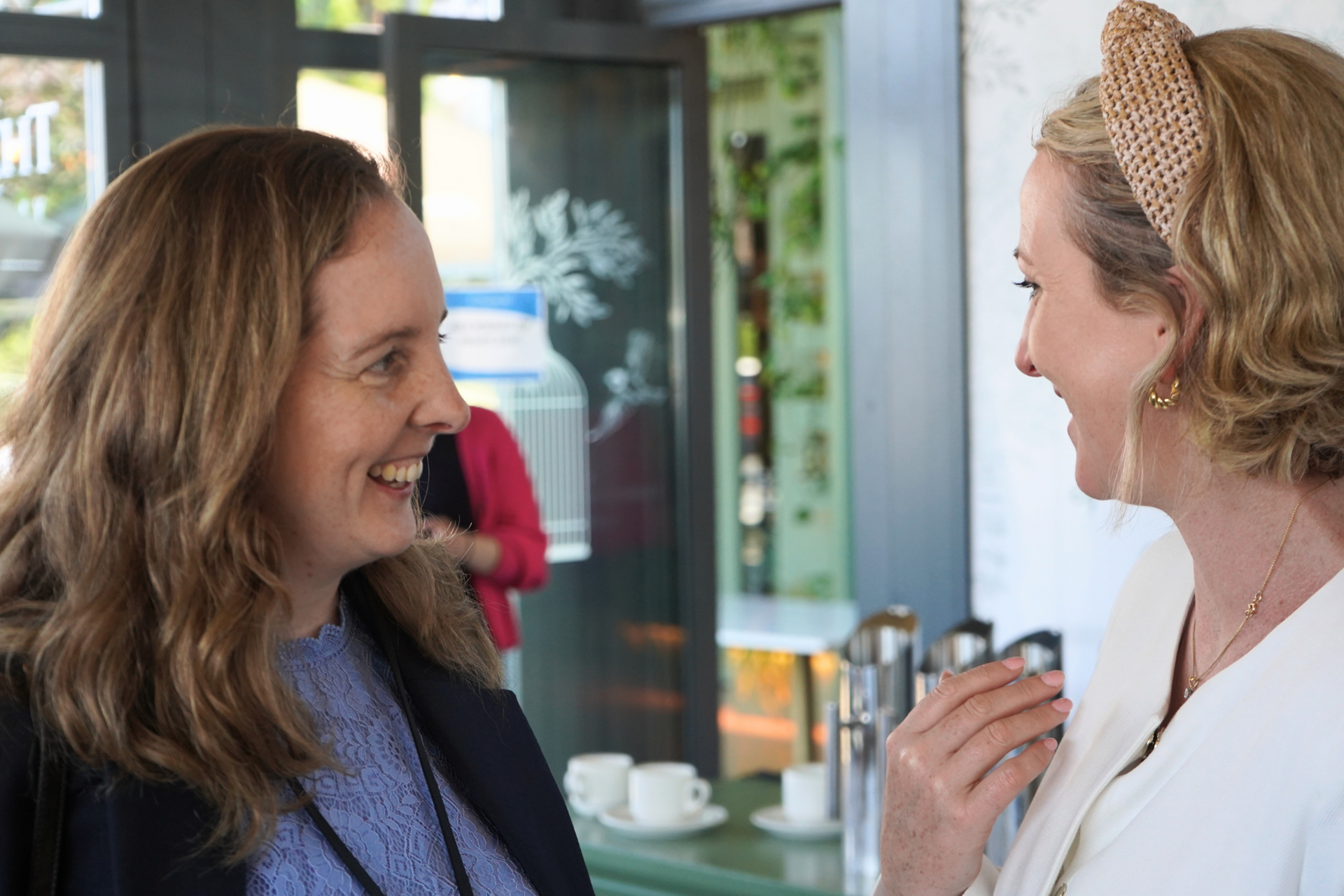 Two women smiling and talking at an indoor event, one holding a drink