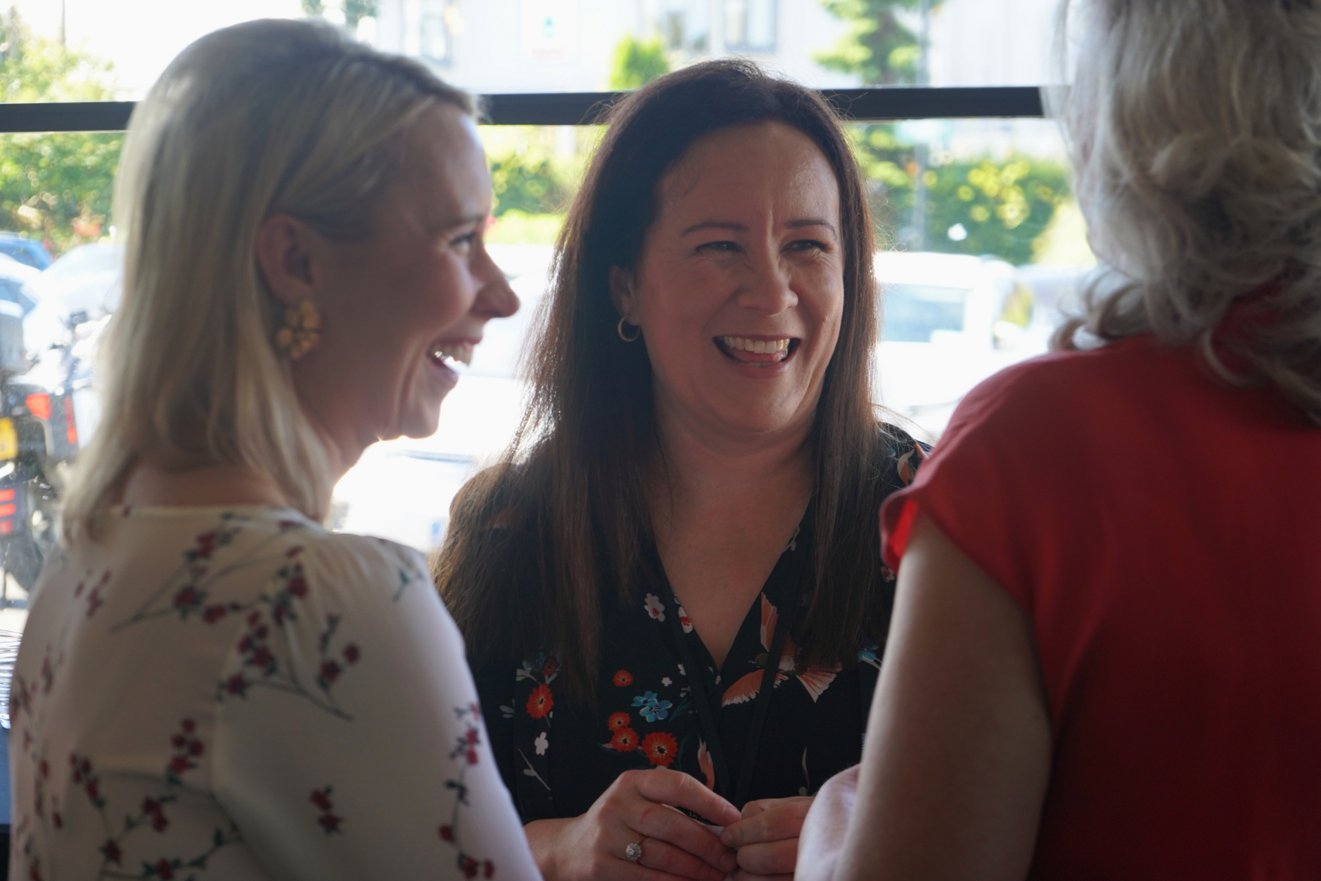 Three women chatting and smiling near a window in a bright indoor setting